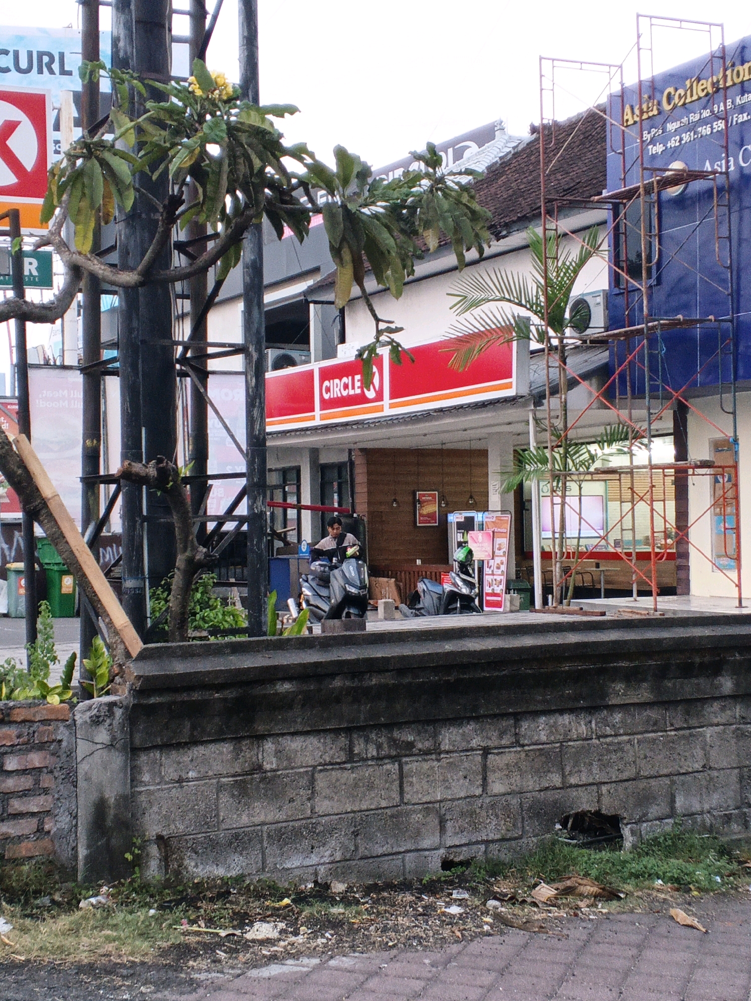 The image shows a street scene in Kuta, Indonesia. In the foreground, a weathered concrete wall with some grass and debris is visible. Behind the wall, a paved area leads to a Circle K convenience store. A man wearing a light-colored shirt and dark pants is seated on a scooter in front of the store, looking to his right. Another scooter is parked to the left of the first. The Circle K store has a bright red awning with its logo. Further back, scaffolding is erected against a blue building with the text "Asia Collection" visible. Palm trees and branches of a leafy plant are scattered throughout the scene, with a yellow flower blooming on one of the branches. The sky appears overcast.