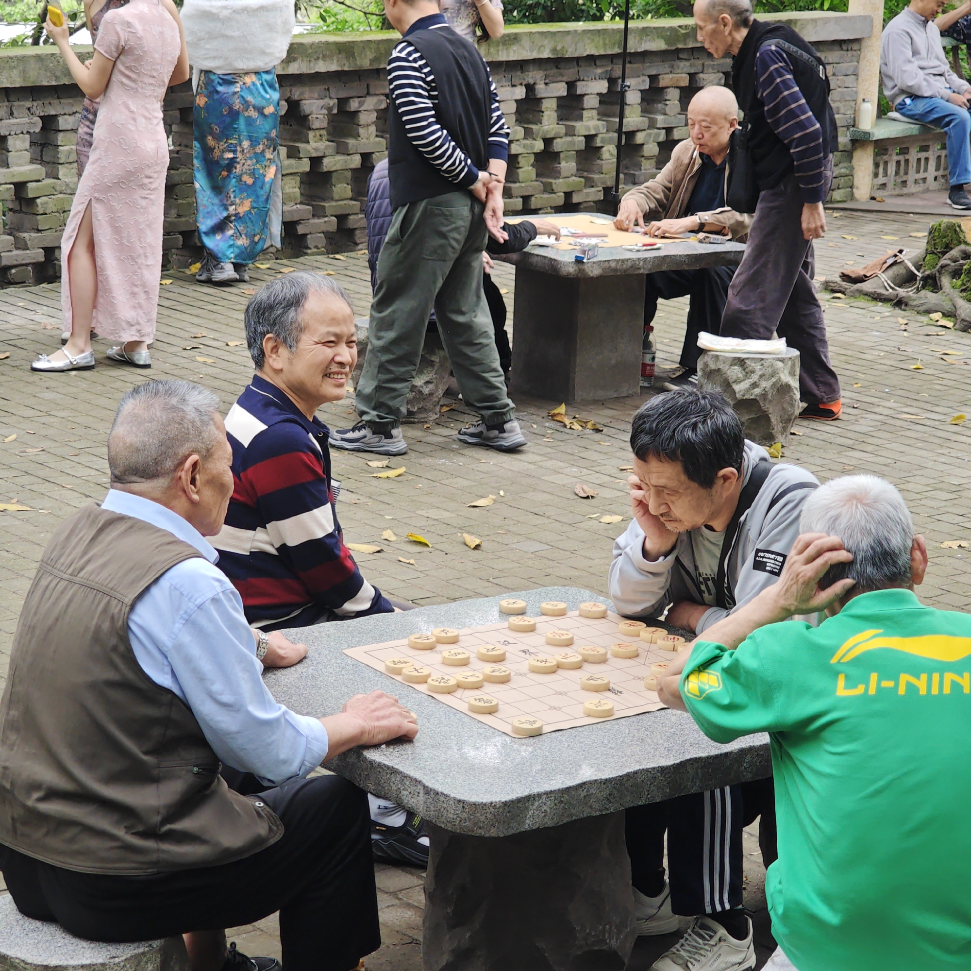 In a public space in Yuzhong District, China, a group of elderly men are engaged in playing Chinese chess (Xiangqi). Four men are gathered around a stone table, with two actively playing the game and two others observing. The chessboard is set up with pieces in play. A man wearing a green "Li-Ning" shirt appears to be contemplating his next move, holding his head, while another man opposite him is also focused on the game. Nearby, at another stone table, more individuals are seen playing cards, observed by others standing. The surrounding area is paved with bricks, and there are other people walking by. The scene suggests a typical leisurely activity taking place in a public park or gathering space.