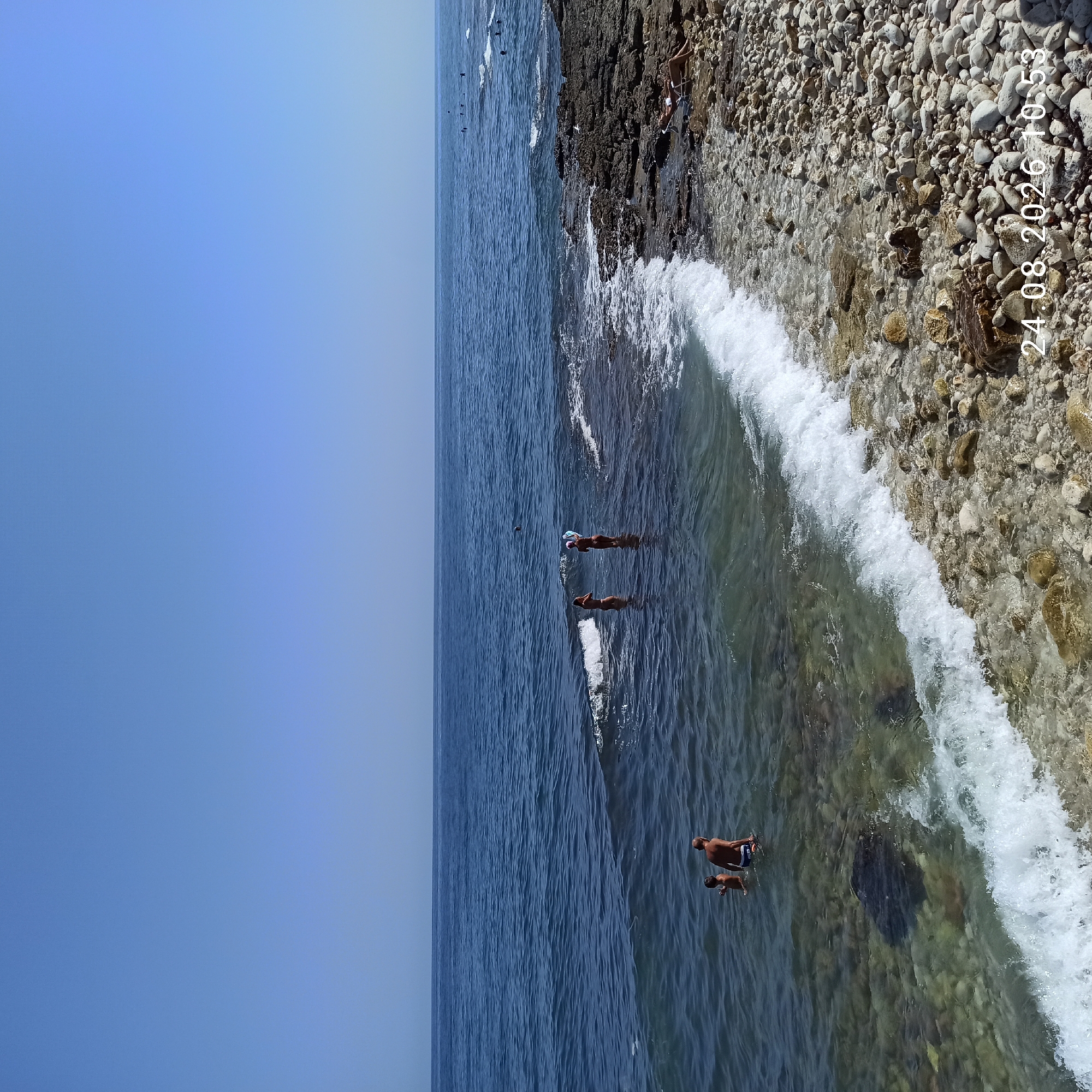 The image depicts a coastal scene on August 24, 2026, at 10:53 AM. The environment is a rocky beach meeting the calm, blue sea under a clear sky. In the shallow water near the shore, several individuals are present. Two adults, one male and one child, are standing in the water. Further out, two women are also standing in the sea, one of whom is holding a surfboard. In the distance, a group of people are visible swimming or wading in the ocean. The foreground is dominated by the rocky shoreline with scattered pebbles and a breaking wave washing onto the beach. A person is seated in a lounge chair on the rocky shore to the right of the image. The overall scene suggests a leisurely day at the beach.