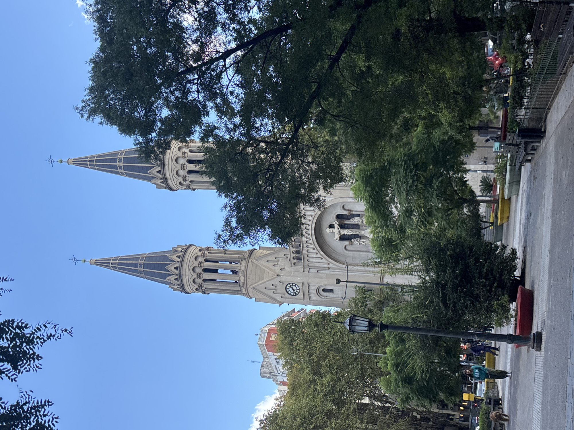 The image displays a grand, ornate stone church with two towering spires, captured on a clear, sunny day. The church features intricate architectural details, including arched windows, decorative carvings, and a visible clock face on one of its spires. Both spires are topped with crosses and dark, patterned conical roofs.
Numerous lush green trees frame the church, with some branches partially obscuring the building and others silhouetted against the bright blue sky. To the right of the church, a paved street or sidewalk is visible, lined with more trees and a traditional black street lamp. Several people are present on the sidewalk; one individual in a light blue/teal top and dark pants is standing, with a dog partially visible nearby. Bright red and yellow planters add splashes of color to the urban environment.
In the background, beyond the trees and street, multi-story buildings are faintly visible. The scene is bathed in bright daylight, indicating a clear and sunny day. The overall impression is that of a bustling urban setting, consistent with a public square or area adjacent to a significant landmark in Buenos Aires, Argentina. No discernible text is visible other than the numbers on the church's clock face.