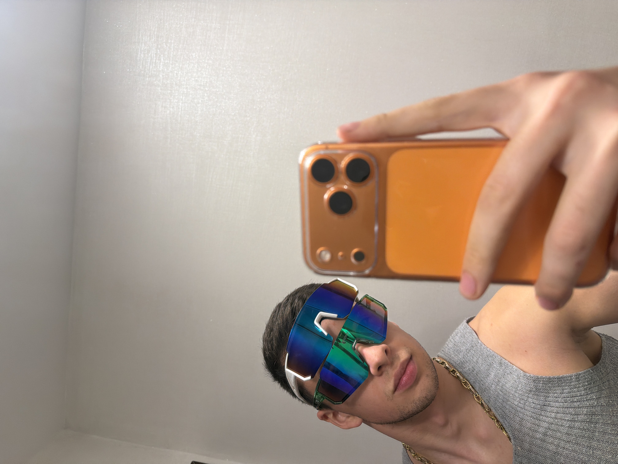 The image captures a selfie taken by a young man, visible from the chest up, positioned towards the bottom center of the frame. He has short dark hair and light stubble, with a neutral expression, his lips slightly parted. He wears a grey ribbed crew-neck top and a prominent gold chain around his neck. His eyes are completely covered by large, sporty-style sunglasses featuring highly reflective, iridescent lenses that display vibrant blue, green, and purple hues. The sunglasses have a white frame.
He is holding an orange smartphone, encased in a clear protective cover, in his right hand. The phone is angled downwards, extending from the right side of the frame towards the top, with his fingers wrapped around it. The back of the phone, showcasing its three distinct camera lenses arranged in a triangular formation on a square module, is clearly visible.
The background is a plain, light-colored wall, likely white or light grey, with a subtle textured finish. A faint corner of the room is visible on the left side of the background, suggesting an indoor setting. The lighting is bright and even, without harsh shadows, indicating good indoor illumination. No specific time of day or precise location can be determined, and there is no visible text in the image.
