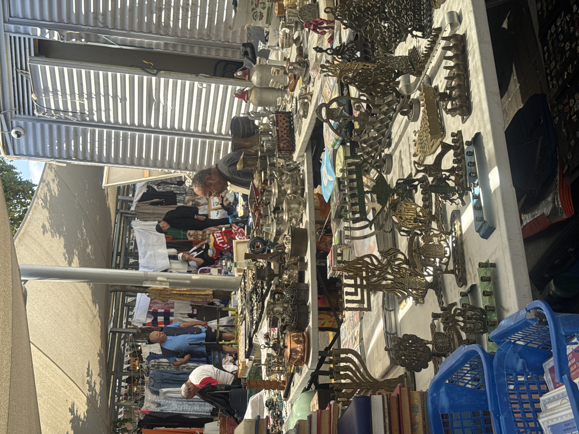 The image depicts a busy scene at a market in Tel Aviv, Israel. The focus is on a stall selling a variety of metal decorative objects, including ornate keys, and other items. A man is examining the merchandise. Other people are visible browsing at nearby stalls, which display clothing and other goods. The market is covered by a corrugated metal awning. Blue plastic baskets are placed behind books at the front of the stall.