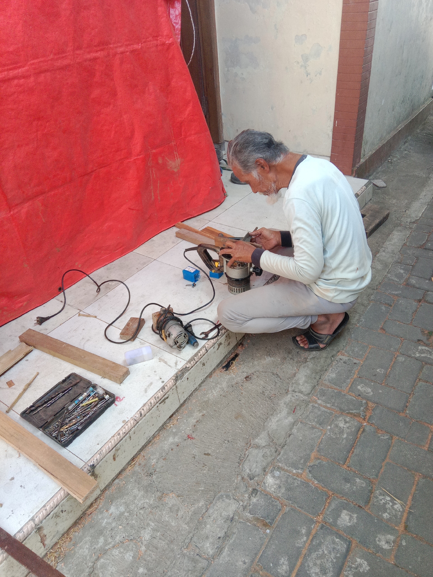 An elderly man with gray hair and a white beard is crouching on a tiled outdoor floor, working on a wooden object with a router. He is wearing a white long-sleeved shirt and light-colored pants, along with black flip-flops. A red tarp is visible behind him, and various woodworking tools, including a router, a drill bit set, wooden planks, and a wrench, are scattered on the floor. The setting appears to be a residential area in Dangkel, Indonesia, with a brick-patterned pavement visible to the right. The overall scene suggests a DIY woodworking project taking place outdoors.