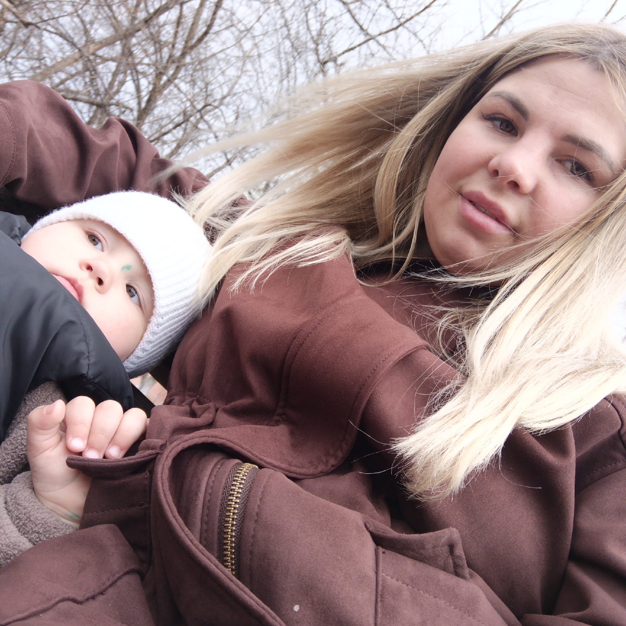 The photograph captures a woman holding a baby against a backdrop of bare tree branches. The woman has blonde hair, and she is wearing a brown jacket. The baby is wearing a white hat and is wrapped in a black covering. A small green mark is visible on the baby's forehead. The location is the city of Unknown, Unknown.