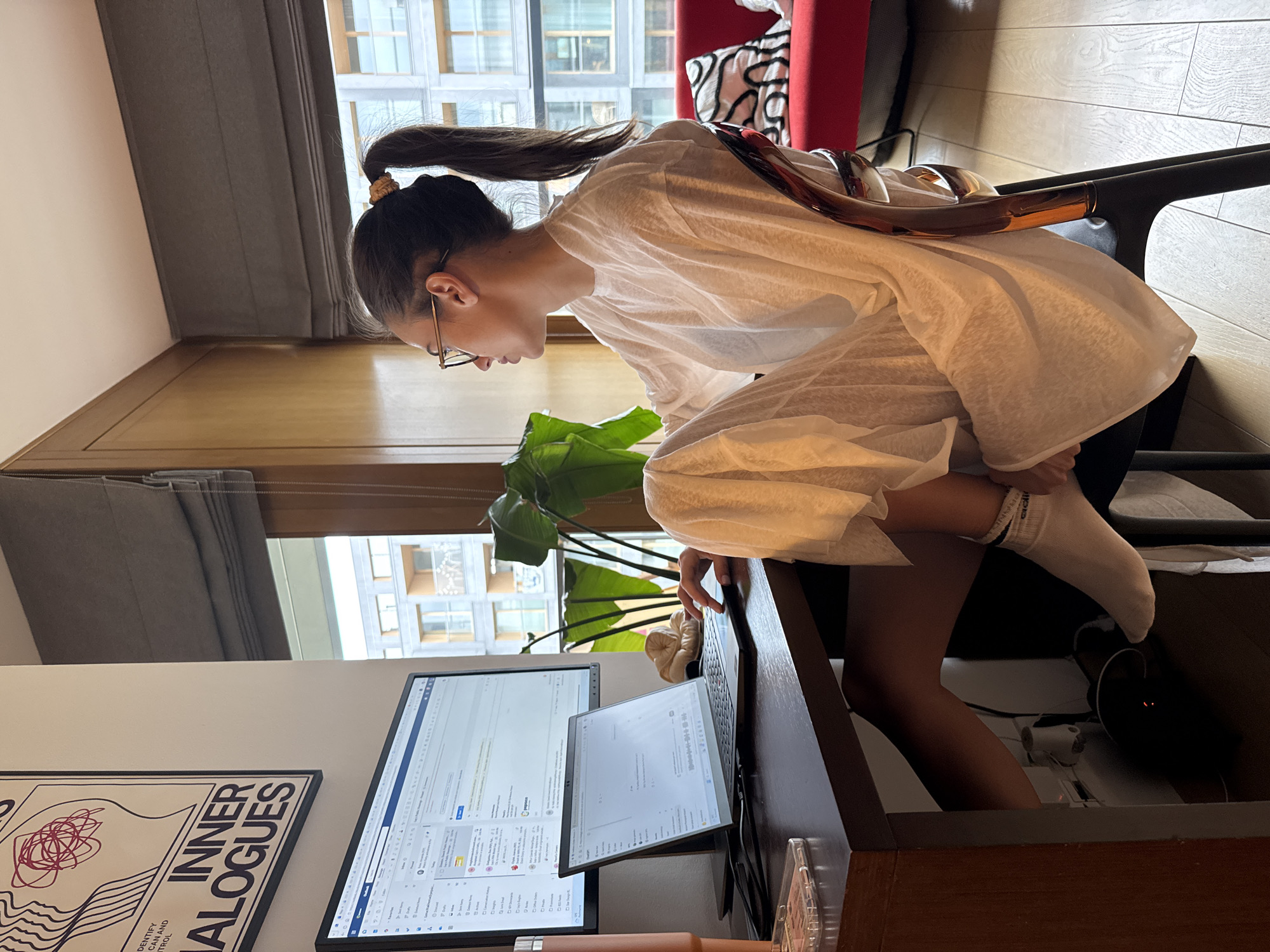 The image shows a young woman sitting at a desk in an indoor environment that appears to be an apartment. She has dark hair pulled back in a ponytail and is wearing glasses and a white long-sleeved shirt. She is leaning forward towards a desk. Her left leg is bent and her foot is resting on the lower portion of the desk structure, and she is wearing a white sock on that foot.
The desk is made of dark wood and holds two computer screens. The larger screen on the left displays what appears to be a spreadsheet application, while the smaller screen on the right shows a document. There is also a laptop on the desk.
Behind the woman, there is a window with a view of a city, featuring buildings in the background. A plant is also visible. The environment seems to be well-lit with natural light coming through the window. There is a poster to the left with the words "INNER DIALOGUES". A red chair can be partially seen behind her. The walls are painted in neutral tones. The city is unidentified.
