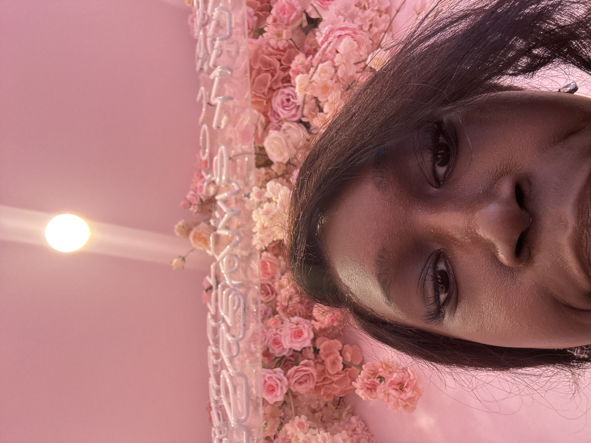 A close-up selfie captures the upper half of a person's face, with their head tilted to the right within the frame. The subject has dark skin and long, dark hair that frames their face, with some strands falling across their forehead. They look directly into the camera with a neutral expression, their eye featuring prominent eyelashes.
The background is predominantly a vibrant pink. On the right side, there is an elaborate floral wall arrangement densely packed with artificial flowers in various shades of pink, rose, and white, creating a soft and romantic aesthetic. Intertwined within this floral display and against the pink wall, sections of a white neon sign are visible. Although partially obscured, fragmented letters spelling out parts of "YOU", "ARE", "THE", and "LIGHT" can be discerned, suggesting a phrase like "YOU ARE THE LIGHT".
To the left of the frame, a bright, circular artificial light source emits a warm glow against the pink background, illuminating the scene. The setting appears to be an indoor space, possibly a cafe, photo studio, or an event venue in Glendale, United States of America, designed with a trendy and aesthetically pleasing decor. The image captures a moment of self-portraiture within this decorated environment.