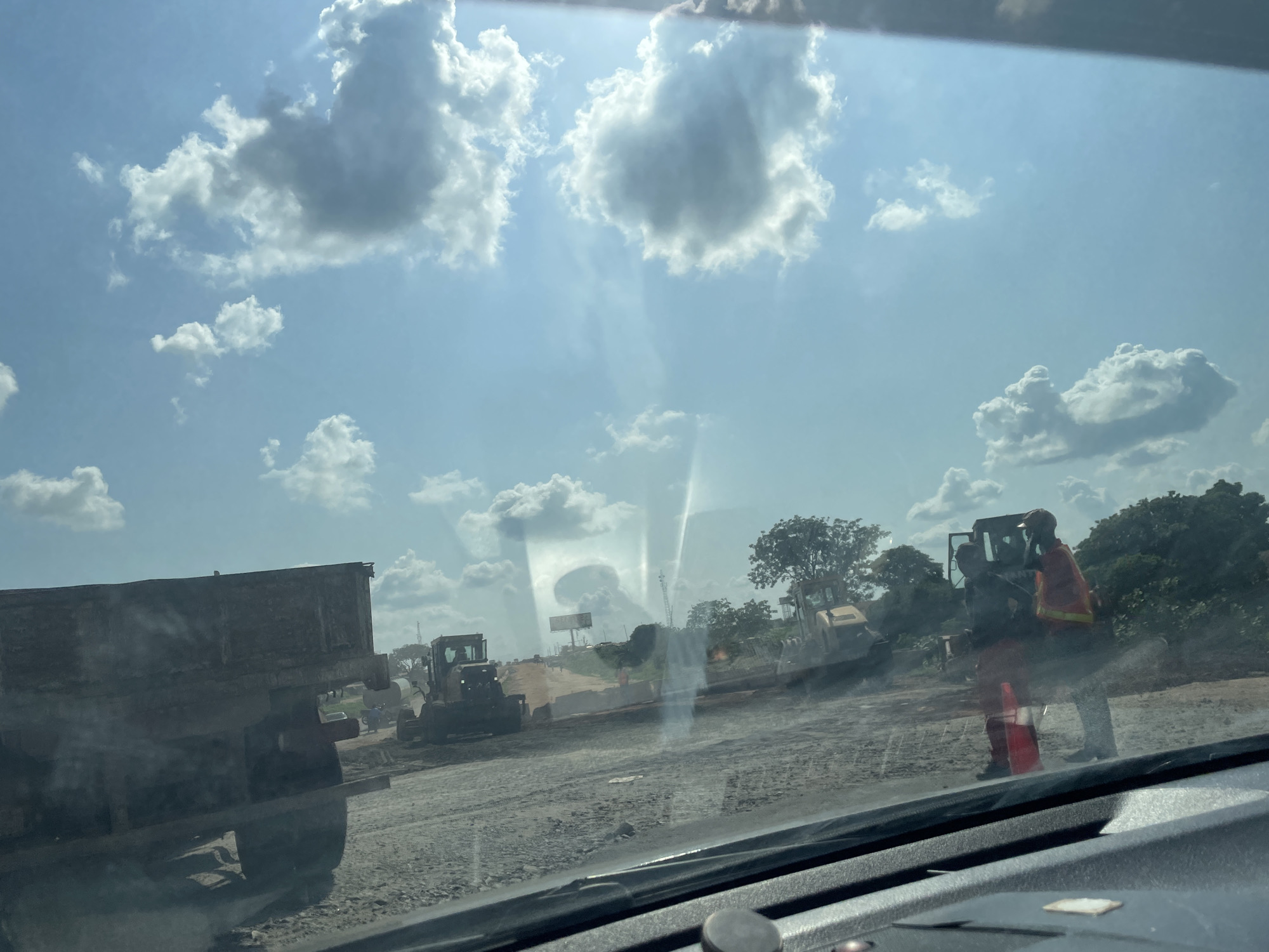 The media file depicts a construction scene under a bright, partly cloudy sky, suggesting it is daytime. The foreground shows the interior of a vehicle, likely a car, with a dashboard visible at the bottom. The view is through a windshield that shows reflections of the sky.
The primary subjects are construction workers and heavy machinery on a dirt road under construction. In the middle ground, there is a large, rusted dump truck. To the right of the dump truck, there are two construction workers interacting. One is wearing a red vest over an orange shirt and appears to be talking to another worker wearing a dark uniform and a red cap. An orange traffic cone is near their feet. Further in the background, a yellow bulldozer is visible, and another piece of construction equipment can be seen to its left. There are trees and vegetation in the distance, as well as what appears to be a billboard or sign. The ground is covered in dirt and gravel, consistent with a road construction site.
The activity taking place is road construction. The presence of heavy machinery and workers indicates ongoing work. The weather appears to be clear with scattered clouds, and the sunlight is strong, creating lens flares on the windshield. The overall impression is a busy work site on a sunny day.
There is no visible text in the image that can be clearly read. The location context is Adonkolo, Nigeria.