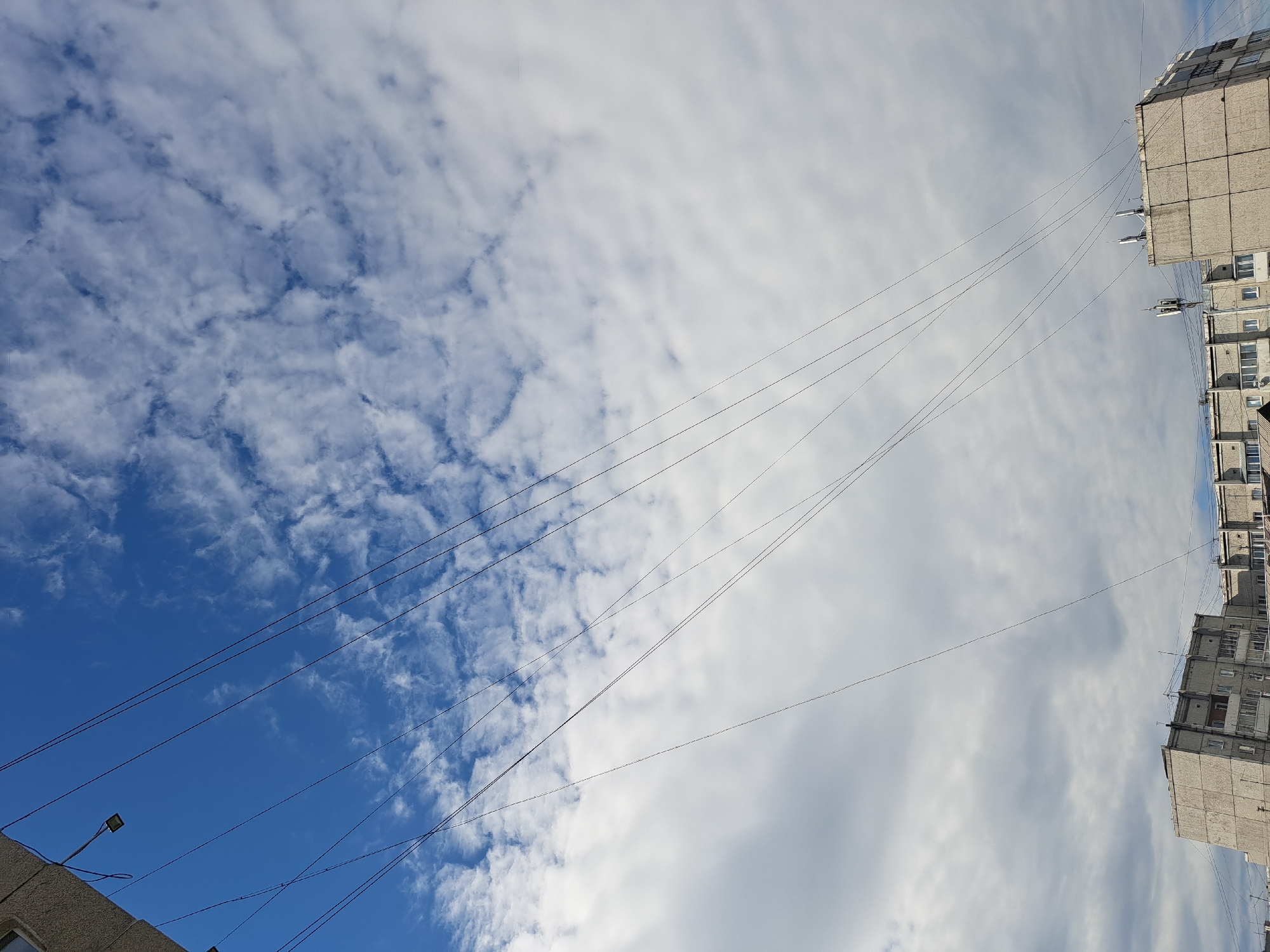 The image captures a daytime scene looking upwards towards a sky filled with clouds against a blue expanse. Several dark, thin wires are diagonally stretched across the frame, originating from the upper left and extending downwards towards the right. In the lower portion of the image, the tops of two grey, multi-story buildings are visible, their concrete facades showing signs of wear. The buildings have numerous windows, and various antennas and equipment are mounted on their roofs. One building on the left appears to be an apartment block, while the one on the right has a more industrial or utility-like appearance. The scene is set in a city environment, likely in Irkutsk, Russia, given the architectural style of the buildings. No people are visible in the image. The lighting suggests it is daytime, with the sun partially obscured by clouds.