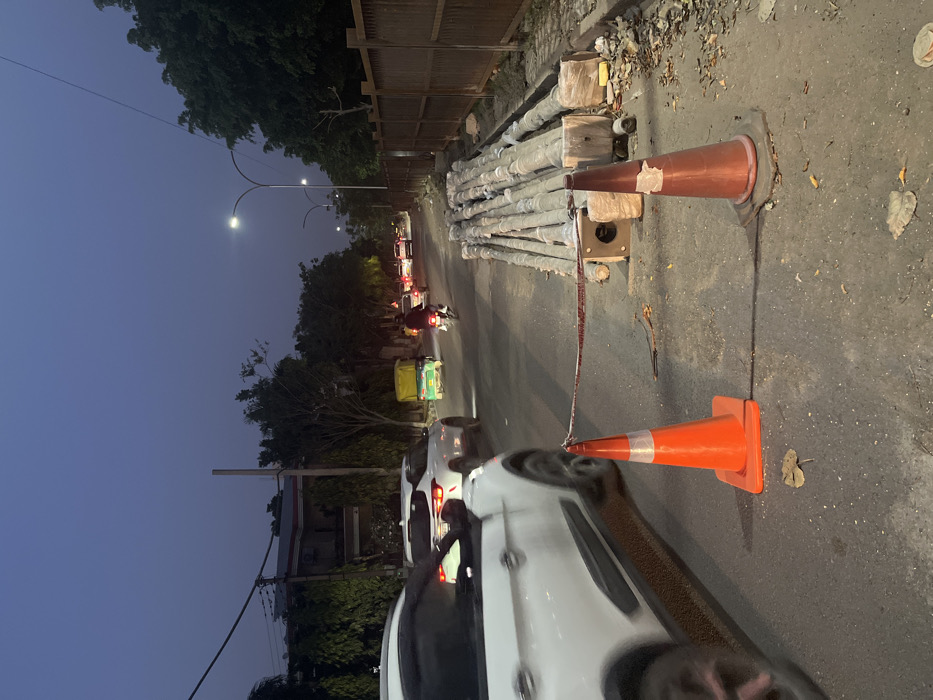 The image shows a street in Sahibzada Ajit Singh Nagar, India, at dusk. A white SUV is stopped on the right side of the street, facing the camera, while a silver SUV drives towards the camera in the left lane. A motorcycle is driving towards the camera in the right lane. A stack of concrete poles is blocking the road. Two traffic cones are placed in the road to warn oncoming traffic about the construction. There is a wooden fence to the right of the road. The sky is dark blue with no clouds. There are trees and houses along the street. The streetlights are on, illuminating the street. There is a man riding a motorcycle in the middle of the street. There are multiple vehicles parked along the right side of the street.