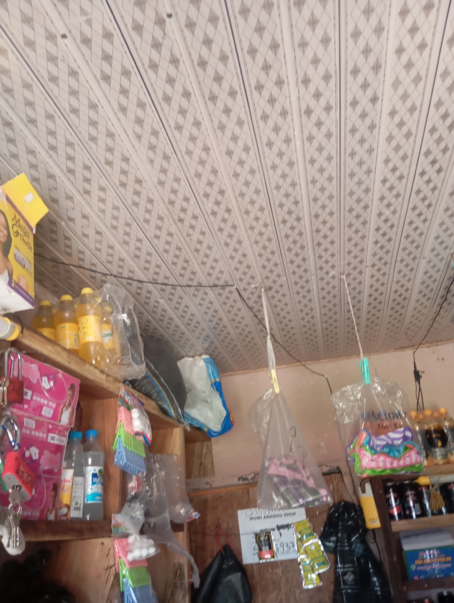 A low-angle, interior shot shows a shelf stocked with various items. On the left, a yellow box labeled "Mega Growth" is visible. Below that, several bottles of yellow liquid are stacked, some in plastic packaging. To the right of these, a blue and white bag rests on a shelf. Further to the right, a clear plastic bag contains colorful items, possibly toys or packaged goods. Below this, a shelf holds several cans, some black and red, and a box with blue and white labeling. The ceiling is paneled with a diamond pattern. A white sign with black text reading "MUMI ARADOU SHOP" is affixed to the wall, with some yellow items beneath it. Several items are hanging from the ceiling via strings and clips. The scene appears to be inside a small shop in Jalingo, Nigeria.