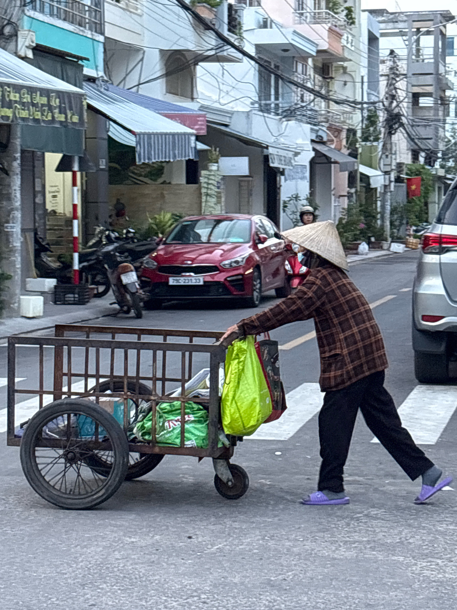 In Nha Trang, Vietnam, a person wearing a conical hat and a plaid shirt pushes a wheeled cart filled with green plastic bags across a street. The cart has a large spoked wheel on the left and a smaller caster wheel on the right. The person is wearing dark pants and purple flip-flops. A red car is visible in the background, and a silver SUV is to the right of the frame. The street appears to be a residential or commercial area, with buildings lining the street and electrical wires overhead. The scene is captured in daylight.