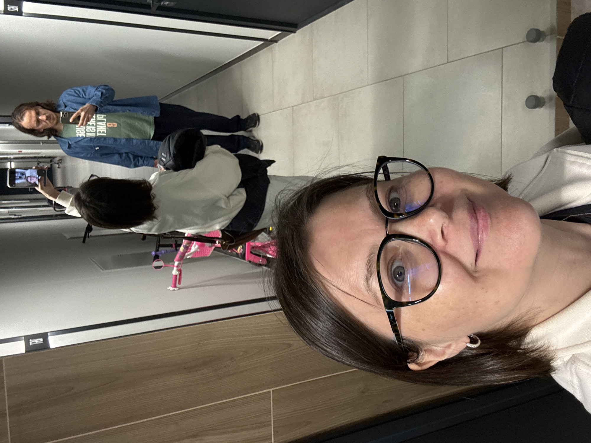 The image captures a complex, layered scene primarily focused on multiple reflections within an indoor setting, likely a hallway or corridor in Izhevsk, Russia. The photograph is oriented vertically, rotated 90 degrees counter-clockwise.
In the foreground, the face and upper torso of a woman are prominently displayed. She wears large, round, dark-framed glasses and a small silver hoop earring in her visible ear. Her dark hair frames a neutral to slightly smiling expression as she looks towards the camera. She is dressed in a light-colored, possibly off-white, hooded top.
Reflected in a mirror behind her, another person is seen from the back, holding a smartphone to capture the scene. This individual is also wearing a light-colored hooded top. A small, pink child's bicycle is visible near their feet on the light-tiled floor.
Further in the reflection, a third individual (or another reflection of one of the previous subjects) stands, visible from the waist up. This person has longer, dark hair and is wearing a blue denim jacket over a green t-shirt. The reversed text on the green t-shirt reads "PLANET B IS NOT AN OPTION". This individual is also holding a smartphone, seemingly taking a mirror selfie.
The background shows light-colored tiled walls, featuring two silver coat hooks, and a floor composed of both light tiles and a section of wooden-patterned flooring. The overall activity involves individuals taking photos with their smartphones, creating a layered effect of reflections. The setting appears casual, under indoor lighting.