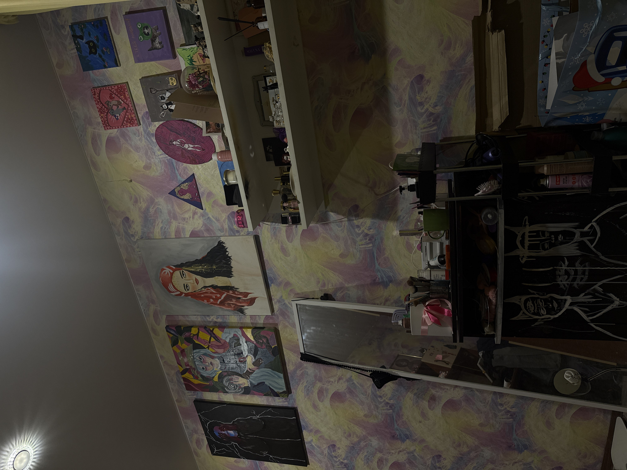 The image depicts an indoor scene, likely a bedroom or studio, characterized by colorful, abstract wallpaper. Several framed paintings are displayed on the wall. From left to right, the first painting is dark with lightning motifs and a figure's face obscured by blue. The second painting features a stylized character with a hat and blue hair. The third painting shows a woman's portrait with a red headscarf.
Above these, on a higher section of the wall, are smaller, square paintings. One shows two cats within a pink heart frame. Another depicts cats in silhouette against a blue background with lily pads. A purple square painting features a cat wearing a frog costume. Below these, a circular pink artwork shows a female figure. A triangular painting with a blonde woman's face is also visible.
A white shelf runs across the wall, holding various items including perfume bottles, jars, and decorative objects. Below this shelf, a tall, rectangular mirror leans against the wall. To the right of the mirror is a black shelving unit containing more items. The lower portion of the image reveals a large, dark painting with white figures that appear to have horns and multiple eyes. To the right of this painting, a large gift bag with a festive Christmas design is visible, along with stacked cardboard boxes.
The lighting appears to be dim, with a ceiling light fixture casting a circular glow in the upper left corner. The overall impression is one of a personalized and artistic living space. The specific location of ЖК «Борисоглебское», Russia, cannot be determined from the visual information provided in the image.