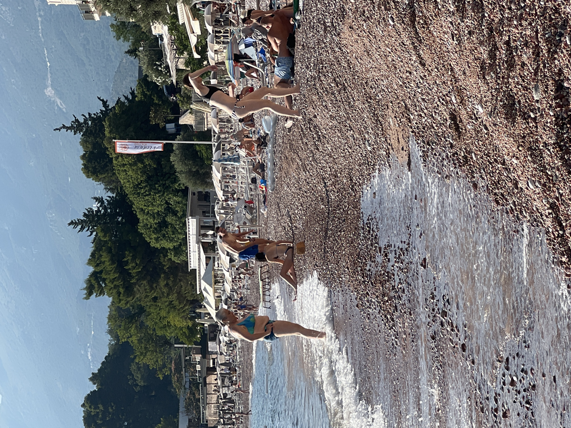 The image depicts a sunny day at a crowded pebble beach, likely in Sveti Stefan, Montenegro. The orientation of the photo is sideways, making it appear as if the beach and sea are vertical. Several people are visible, dressed in swimwear, enjoying the beach. Some are lounging on deck chairs under umbrellas, while others are wading in the shallow water. One woman in the foreground, wearing a blue and green bikini and blue shorts, stands in the gentle surf, her legs submerged. Further back, a man and a woman, both in swimwear, are also near the water's edge. In the mid-ground, a woman in a black bikini stands, possibly posing or adjusting her attire. The beach is lined with rows of white umbrellas and deck chairs, suggesting a commercial establishment. Behind the beach, lush green trees and the silhouette of mountains are visible against a clear blue sky. A white banner with red lettering is attached to a flagpole, but the text is partially obscured and difficult to read clearly. The overall scene conveys a relaxed and leisurely atmosphere characteristic of a summer vacation.