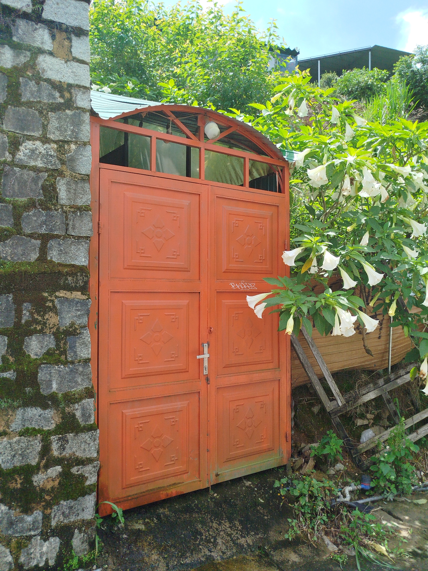 An orange metal double gate stands open to reveal a pathway. The gate is ornate with embossed geometric patterns on each panel. The left side of the image is a rough stone wall covered in moss, and the right side shows a lush green bush with white bell-shaped flowers. Behind the gate, a corrugated metal roof is visible, supported by an orange metal frame. The background features a hillside covered in trees and vegetation, with a dark-tiled roof visible at the top of the hill. The ground in front of the gate appears to be a concrete surface with some debris and scattered vegetation. The overall impression is of an outdoor scene in a somewhat overgrown, possibly rural or semi-rural setting. The location is identified as Xuan Truong - Da Lat, Vietnam.
