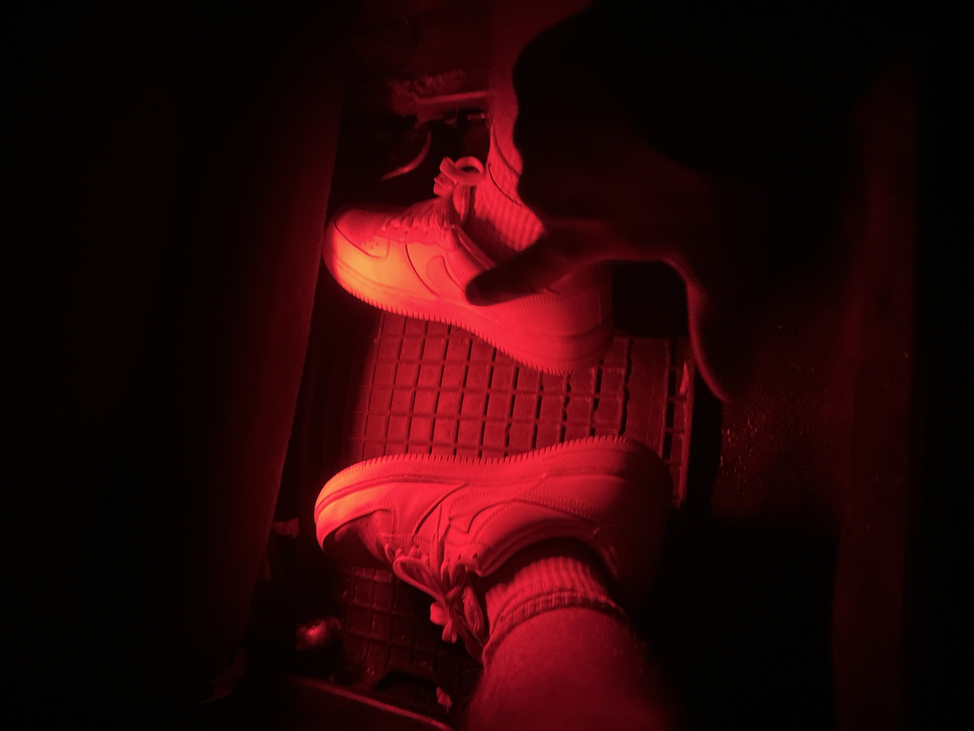 The image shows a close-up view of two feet wearing white sneakers and striped socks, illuminated by red light, suggesting an interior setting possibly within a vehicle due to the presence of pedals and a textured floor mat. One foot is positioned over what appears to be a brake pedal, while the other is near it. A hand with a finger pointed is visible in the foreground, partially obscuring the right foot. The lighting creates a dark, atmospheric environment. The location in Zlatograd, Bulgaria, is not discernible from the visual information provided in the image.