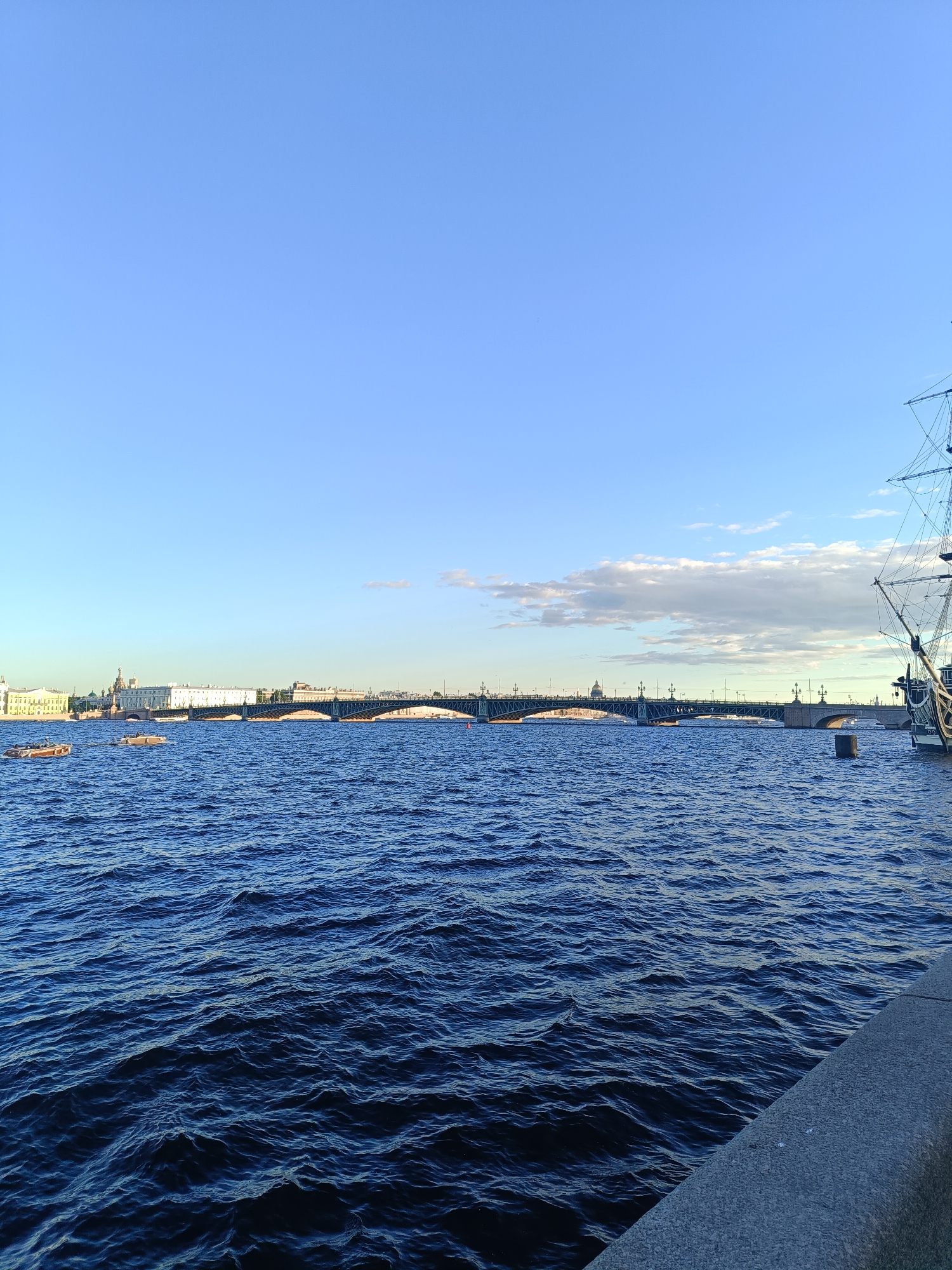 The image shows a wide river in Saint Petersburg, Russia, with a long bridge spanning across it. The water is a deep blue with small waves. In the foreground, a concrete embankment runs along the right side of the frame. A tall sailing ship is docked on the right side of the image. In the distance, across the river, is a cityscape with buildings and a large dome, likely a prominent landmark. Several small boats are visible on the water. The sky is a clear blue with a few scattered clouds.