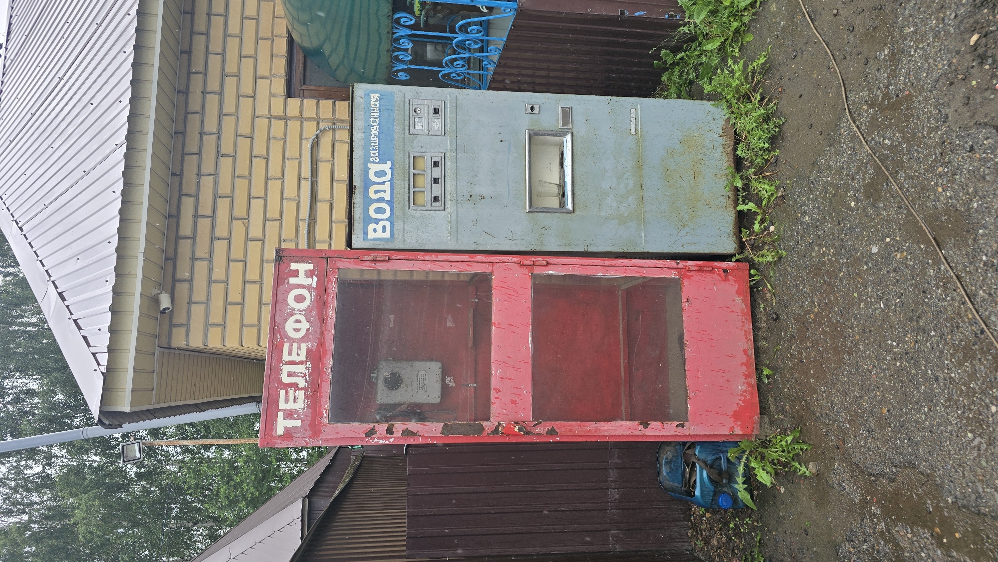 This image captures an outdoor scene in the village of Bol'shoi Mashlyak, Russia, featuring two distinct vending machines situated near a building. On the left is a weathered red telephone booth, its glass panels revealing an old rotary dial phone mounted inside. The booth shows signs of age, with peeling paint and visible wear. To its right stands a pale blue vending machine, labeled in Russian with "ВОДА" (Water) and "газированная" (sparkling). This machine has a dispenser slot with a glass cup inside, and a control panel with buttons and small displays. Both machines are positioned on a gravel surface, with some weeds and greenery growing around their bases. The background includes a building with yellow brickwork and a corrugated metal roof, as well as a dark brown metal fence. The overall impression is of an older, possibly disused, public utility area.