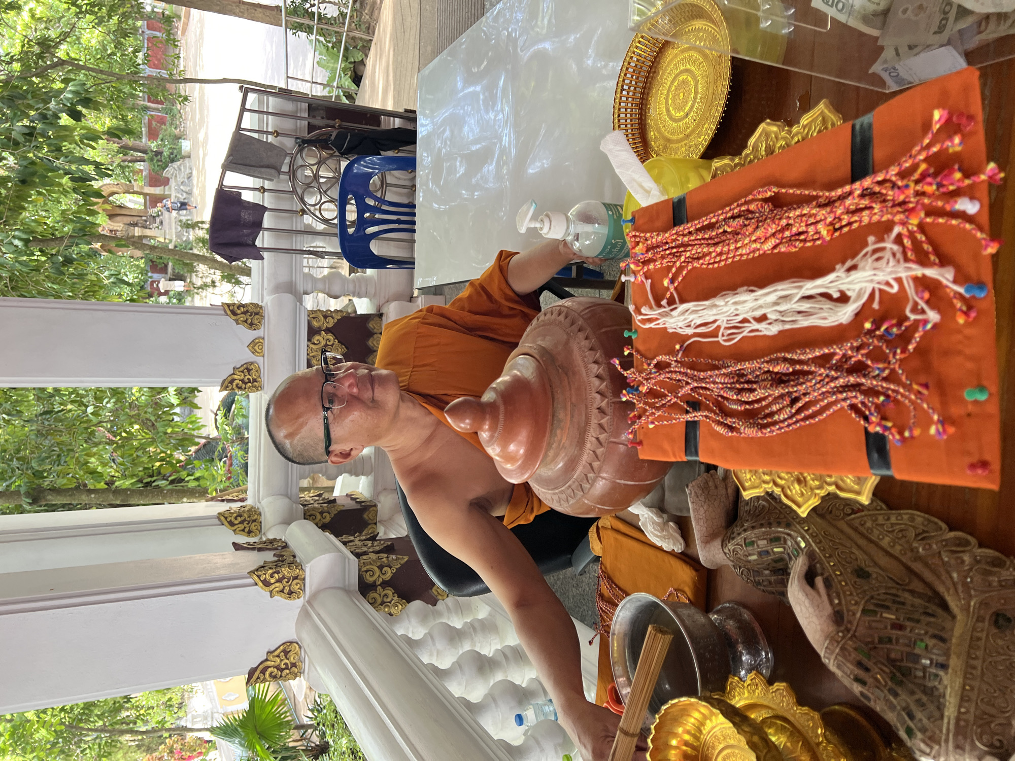 In Baan Hua Thanon, Thailand, a Buddhist monk wearing an orange robe and glasses is seated at a table covered with a white sheet. The monk is reaching towards a metallic bowl containing water and wooden sticks. On the table are several items, including a large terracotta pot, an orange cloth with multi-colored strings attached, a golden tray, and a statue of a reclining figure. A bottle of hand sanitizer is also visible. The scene takes place outdoors, with a white balustrade and lush green trees in the background.
