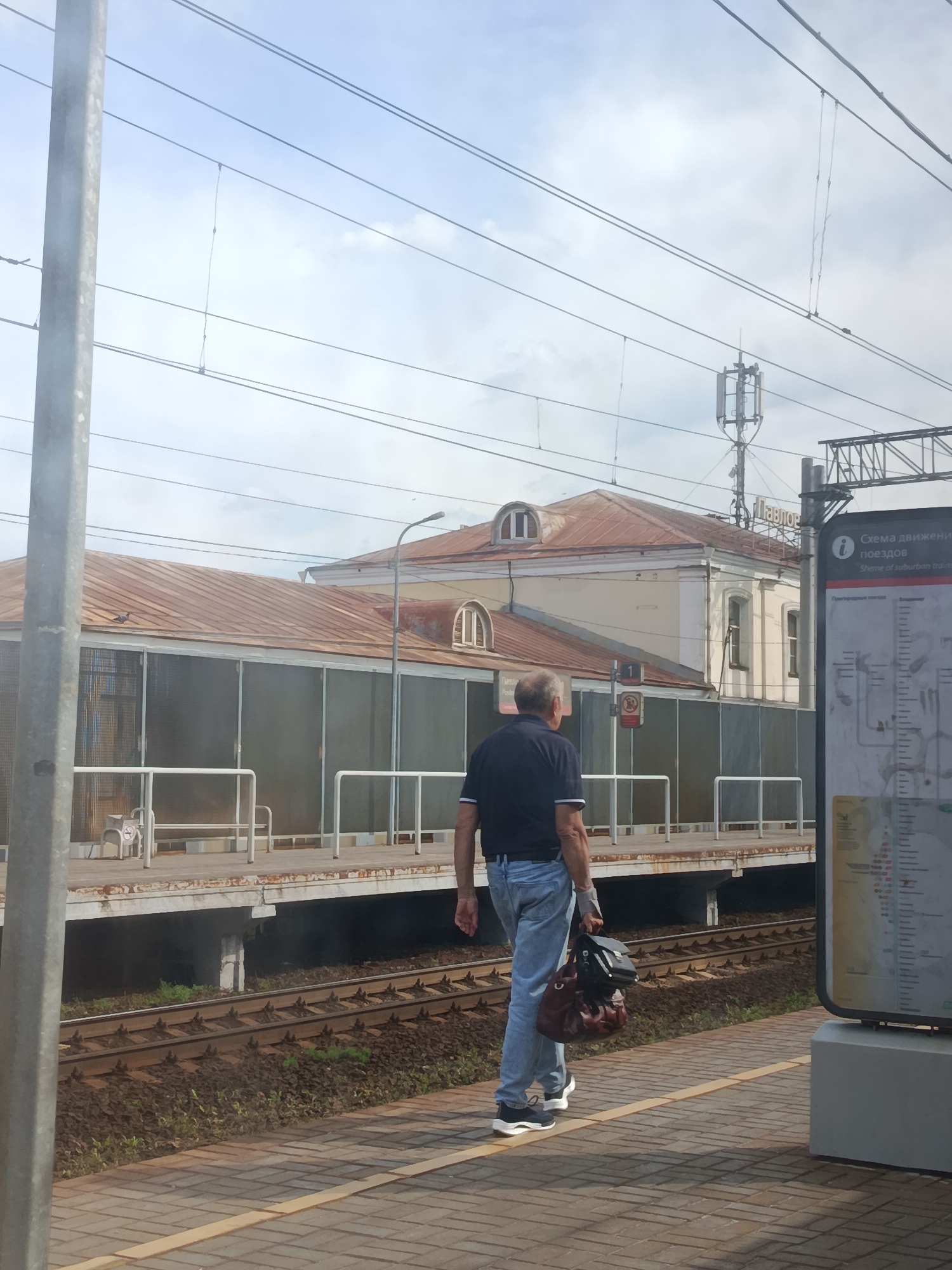 The image shows a man walking on a train platform in Pavlovsky Posad, Russia, carrying two bags. He is wearing a dark blue polo shirt and light blue jeans, and white sneakers. The platform has a yellow line marking its edge. Behind the platform are train tracks and lush green vegetation. Further back, there is a building with a rusty corrugated metal roof, and a smaller building with a light-colored facade and arched windows. Overhead, there are power lines and a cellular tower. A large informational sign is visible on the right side of the image, displaying a diagram of train routes. The sky is overcast.