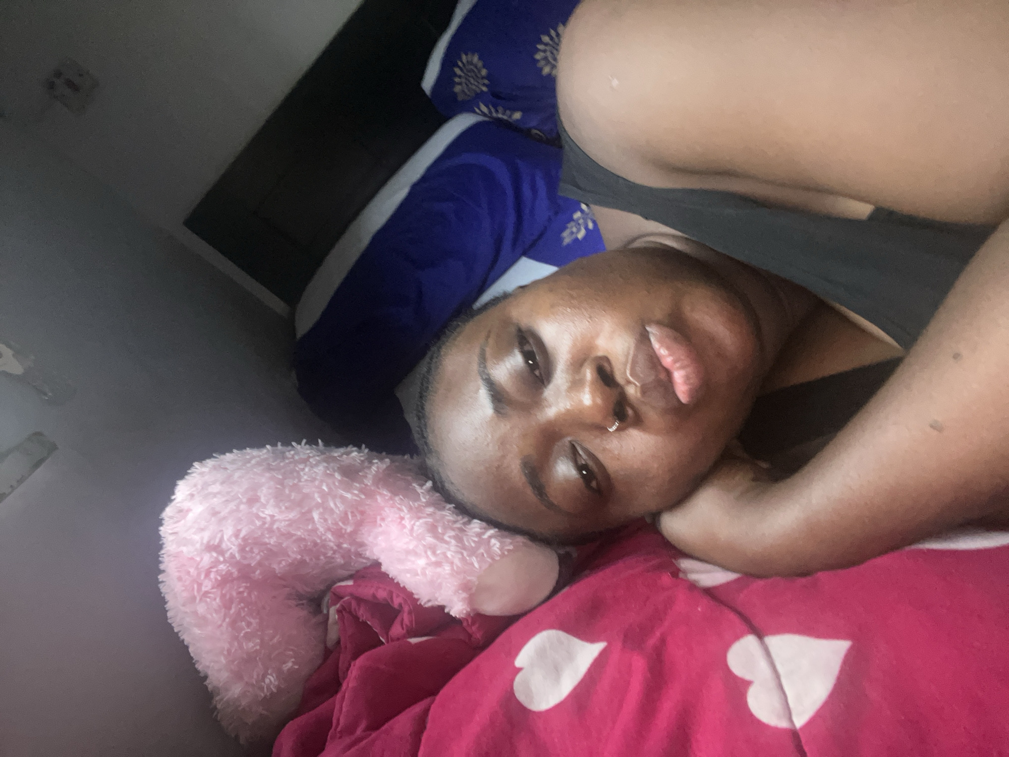 A close-up, slightly tilted shot captures a person lying on a bed in what appears to be a bedroom. The individual has dark skin, a prominent nose piercing, and full lips with a slightly pouting expression. Their eyes are open, looking directly towards the camera, conveying a neutral or relaxed demeanor.
The person is resting their head on a blue pillow featuring a gold or yellow floral pattern. A pink comforter, adorned with distinct white heart shapes, covers the lower part of the frame and the person's body. To the left of the individual, a fluffy, light pink stuffed animal, possibly a teddy bear, is visible. The background shows a dark headboard or wall section, and above it, a lighter-colored wall or ceiling, appearing somewhat dim.
The lighting in the room is low, suggesting it might be evening or a dimly lit indoor space. The scene evokes a sense of comfort and repose. There is no visible text within the image. The setting is an indoor residential space, with the contextual location being Abuja, Nigeria.