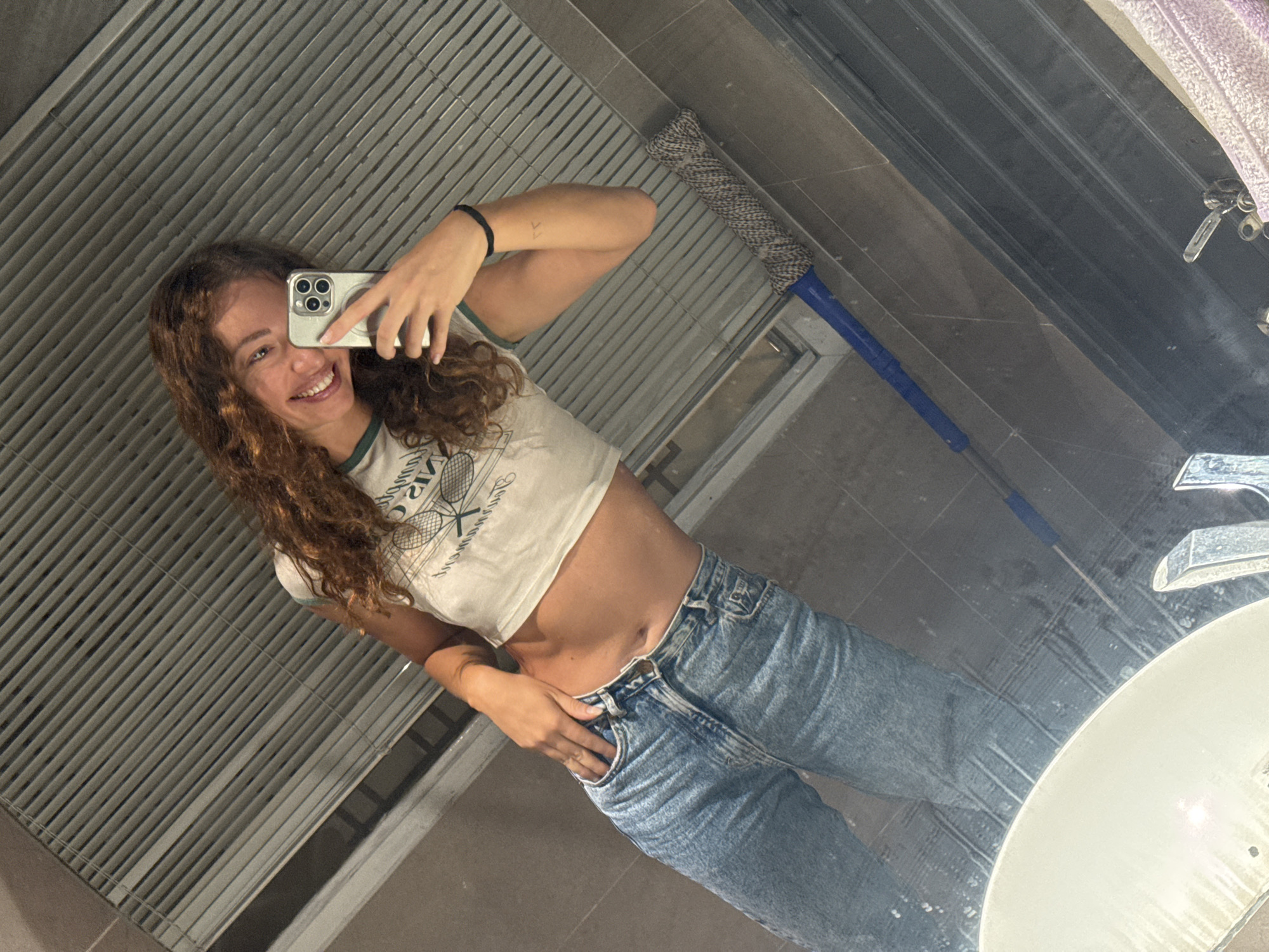 A young woman with curly brown hair, wearing a white crop top and blue jeans, smiles while taking a mirror selfie. She is standing in what appears to be a bathroom. In the reflection, a mop with a blue handle and a grey head is visible, leaning against a tiled wall. The background features a grey, slatted ceiling or ventilation grate. The overall scene is casual and candid. This image is not in Hà Nội, Vietnam.
