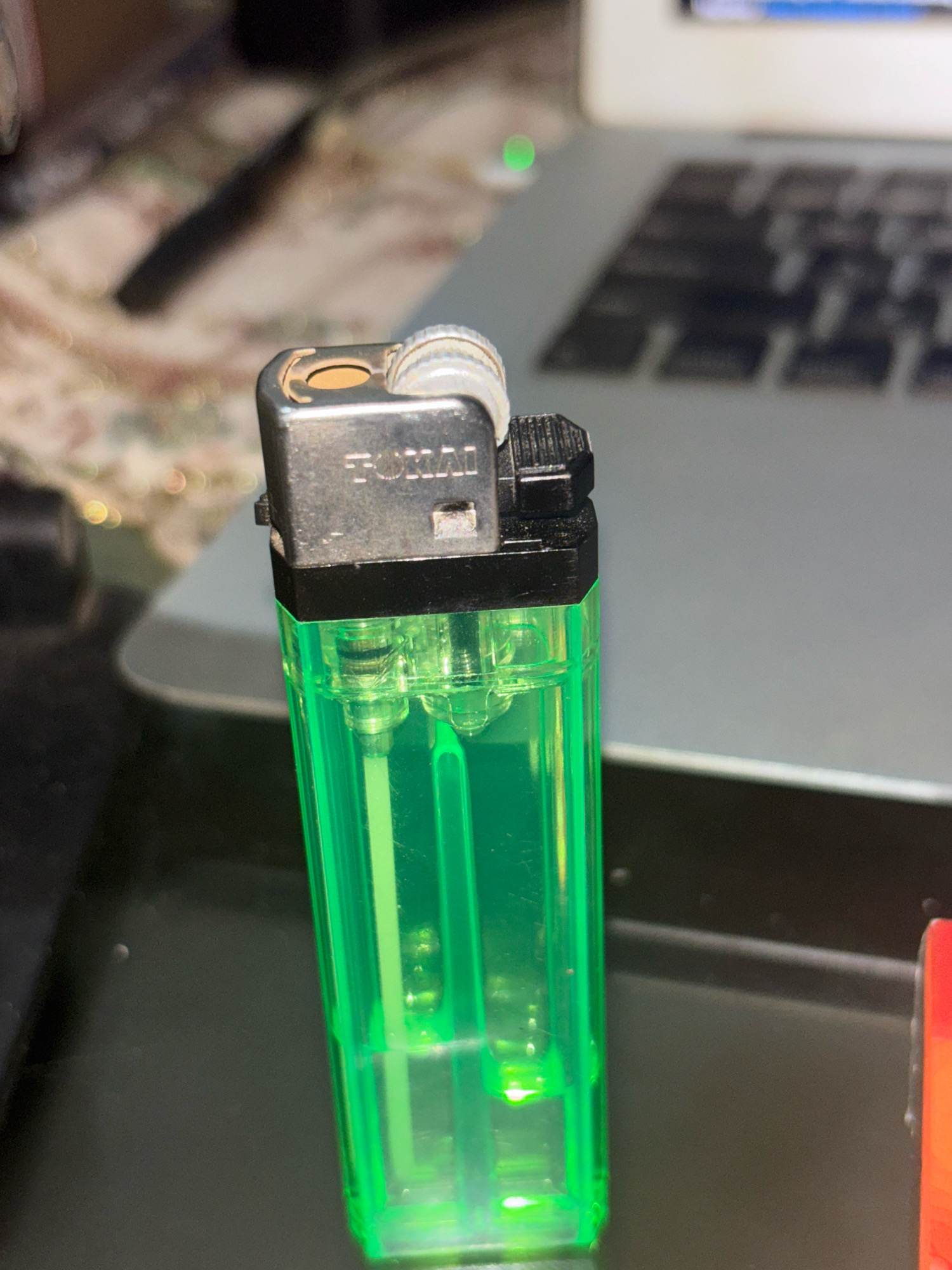 A close-up, slightly high-angle shot focuses on a green disposable lighter. The lighter has a silver metal top with "TOKAI" imprinted on it. The lighter is positioned in the foreground, with its transparent green body filled with liquid fuel visible. Behind the lighter, a laptop keyboard is slightly out of focus, with a green indicator light visible on the laptop's surface. The background is blurred, with hints of patterned fabric and dark surfaces. The scene appears to be indoors, possibly on a desk or table, with no indication of the city of Bekasi, Indonesia.