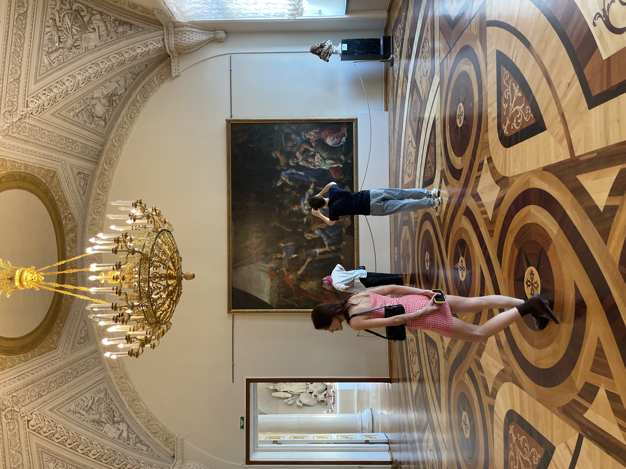 Here is a detailed description of the image:
**Scene Overview:**
The image captures a scene inside a grand, classical-style room that appears to be a part of a museum or an ornate building, possibly located in the city of Unknown, Unknown. The room features elaborate architectural details, including a decorative ceiling with intricate plasterwork, a large painting on the wall, and a beautiful patterned wooden floor. The overall atmosphere is one of opulence and historical significance.
**People:**
There are three individuals within the scene. A man and a woman are standing in front of a large painting looking up at it, the woman is holding her phone and wearing a red and white dress. There is another person standing next to them, and a fourth figure appears in the doorway.
**Objects:**
* **Chandelier:** A large, ornate golden chandelier hangs from the ceiling.
* **Painting:** A large, framed painting is mounted on the wall.
* **Flooring:** The floor is elaborately patterned with inlaid wood.
* **Bust:** A classical bust is placed on a pedestal against the wall.
* **Doorway:** An open doorway reveals another room, suggesting a continuation of the museum or building.
**Actions & Interactions:**
The individuals are primarily focused on observing the art within the room. The man and woman are looking at the painting. The other person standing next to them appears to be either viewing the artwork as well or preparing to do so.
**Environment:**
* **Lighting:** The room is well-lit, with natural light coming from a window.
* **Architecture:** The classical architectural style is evident through the archways, detailed ceiling, and overall proportions of the room.
The scene presents a typical museum-like environment where visitors are appreciating and engaging with historical art and architecture.