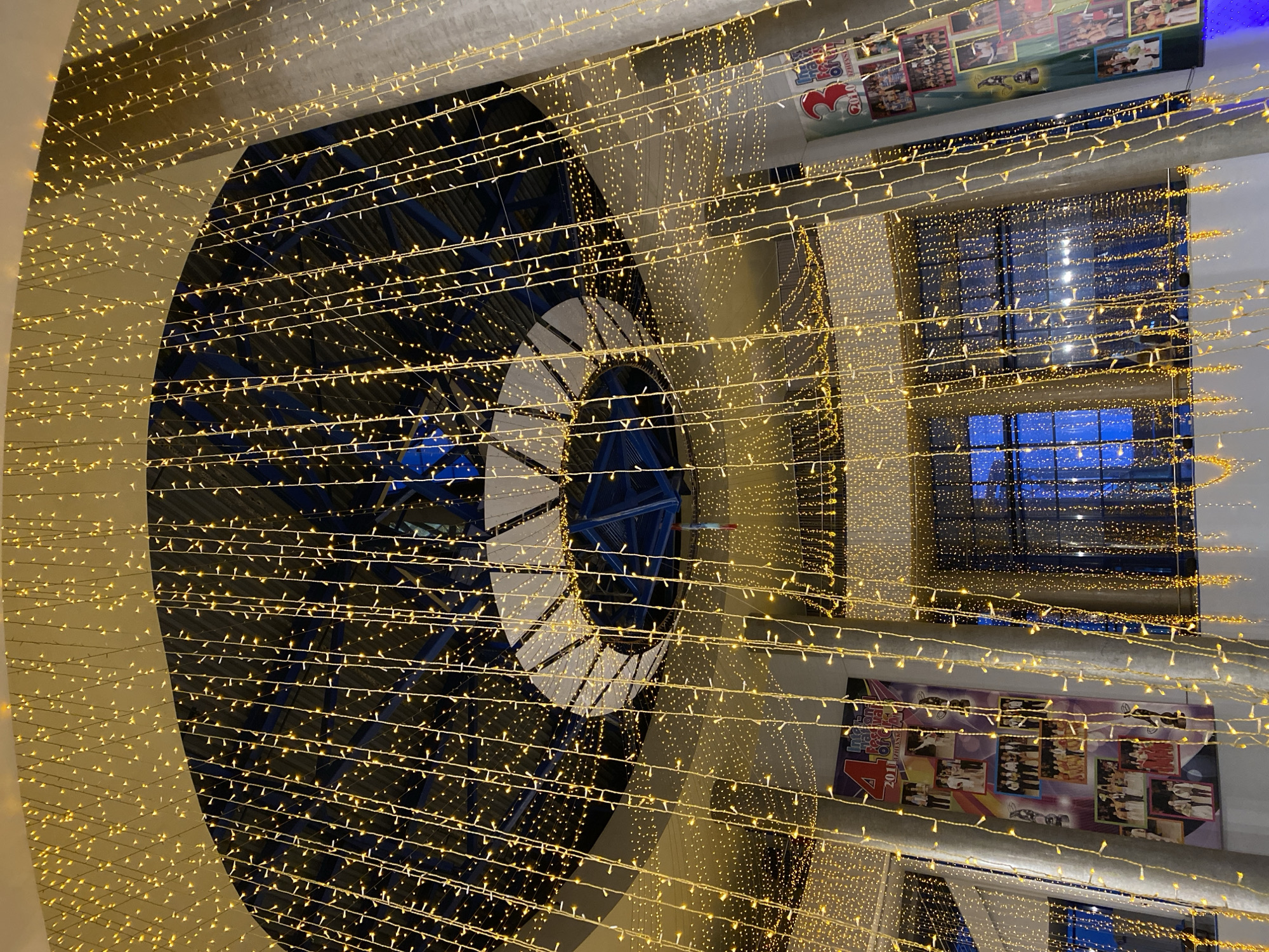 This indoor image, taken in Izhevsk, Russia, shows a complex architectural element adorned with strings of small yellow lights. The primary element is a large, circular structure with a blue and white color scheme, possibly part of a building's internal design or decoration. The lights are draped in numerous horizontal strands, creating a luminous effect. The background includes a building's interior, with windows revealing a blue, possibly evening or nighttime sky. Posters or decorative panels are visible on the walls. The composition is dynamic, with lines and geometric shapes created by the lights and the architectural elements. The lack of people suggests the image was captured in a quiet moment or off-peak hours.