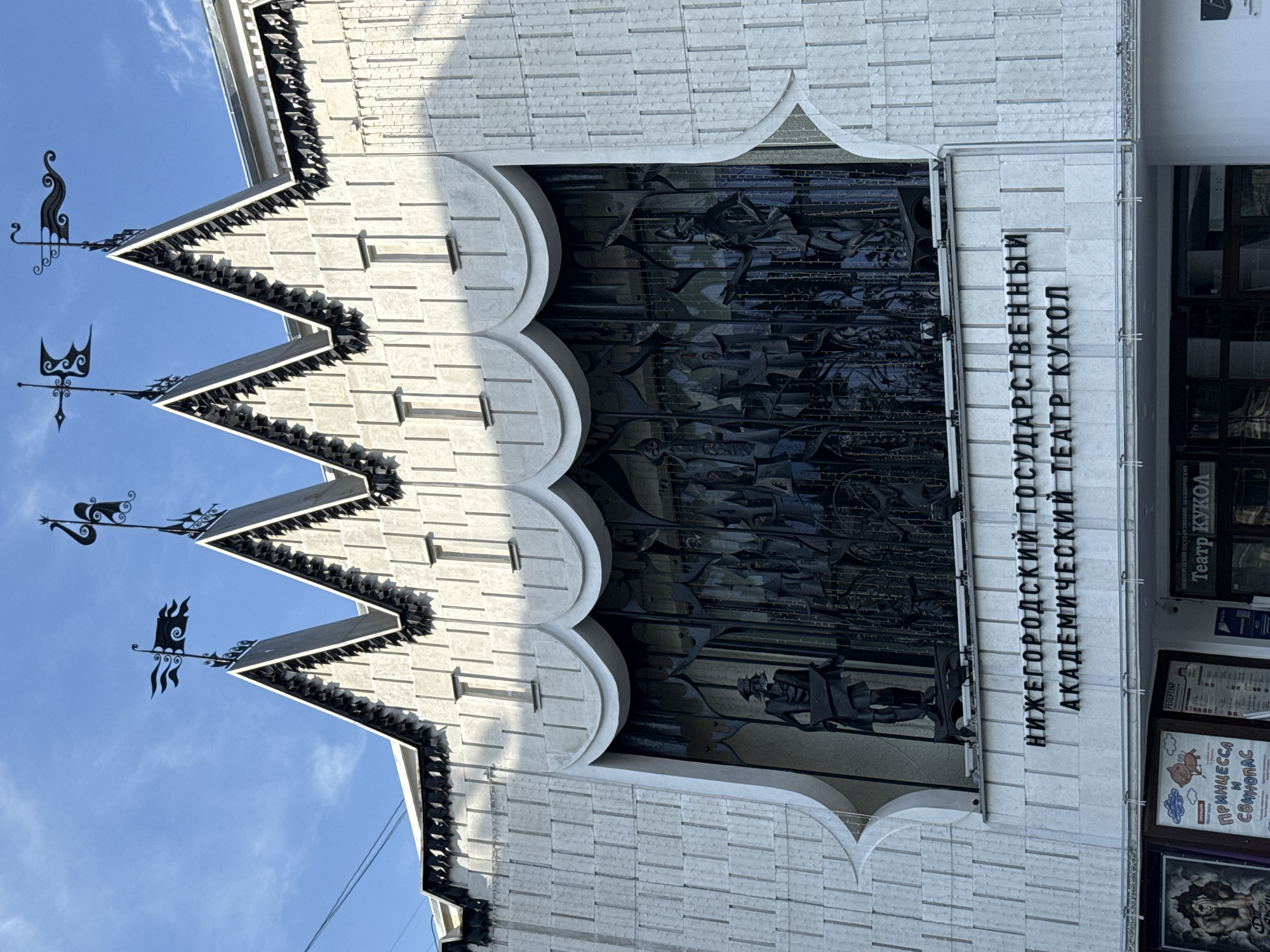 The image shows the exterior of the Nizhny Novgorod State Academic Puppet Theatre. The building's facade is predominantly white with a textured finish, featuring a series of pointed, decorative roof elements topped with black metal weathervanes. A large, ornate black metal screen covers a significant portion of the facade, depicting figures that appear to be from a performance. Below this screen, in raised black lettering on a white marble surface, are the words "НИЖЕГОРОДСКИЙ ГОСУДАРСТВЕННЫЙ АКАДЕМИЧЕСКИЙ ТЕАТР КУКОЛ" (Nizhny Novgorod State Academic Puppet Theatre). At the very bottom of the visible facade, there are posters and entrances to the theatre. The sky above is a clear blue with a few wisps of clouds. The lighting suggests it is daytime, with shadows cast by the building's architectural features.