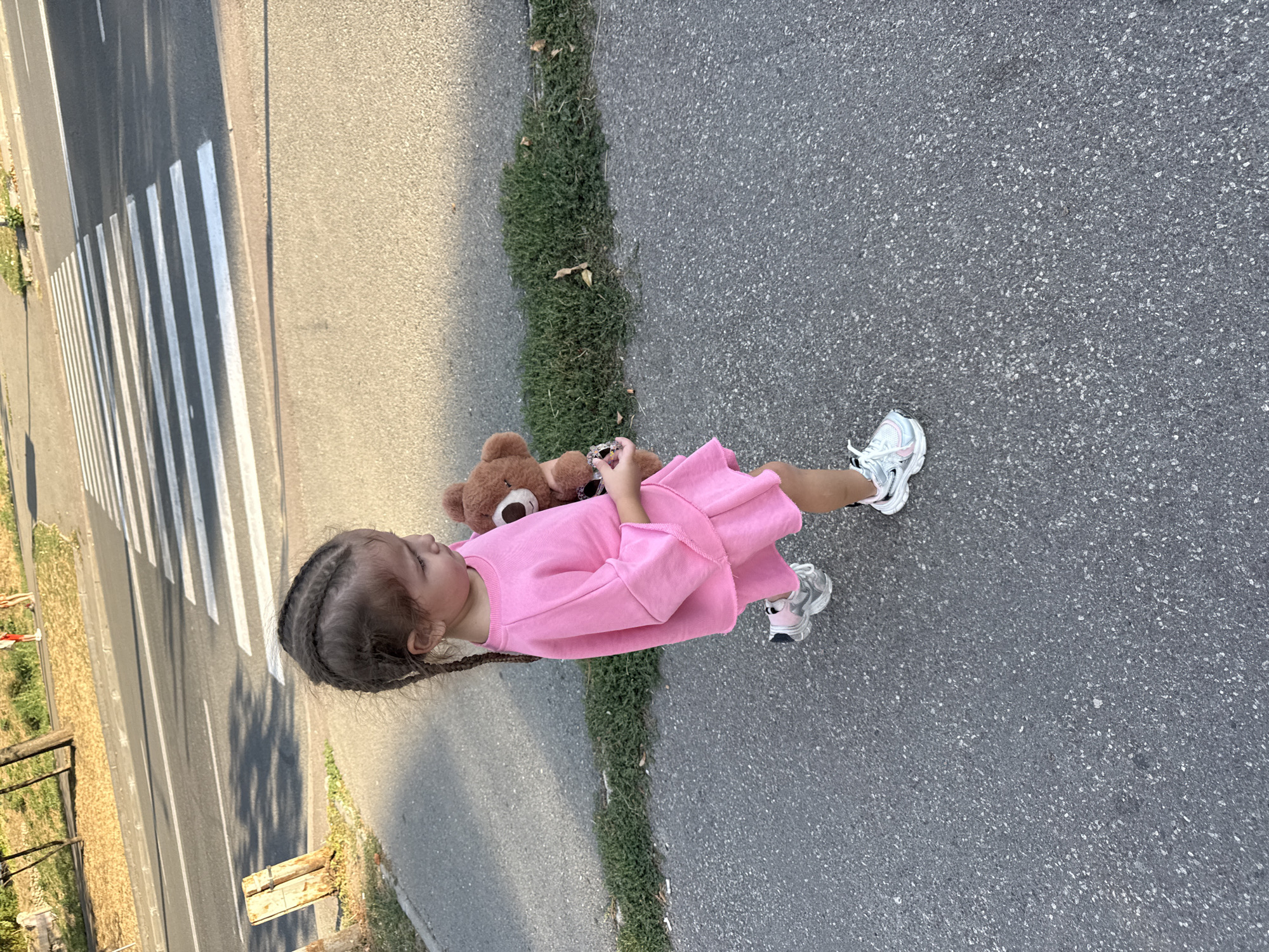 A young girl with braided hair walks on an asphalt road, carrying a brown stuffed bear. She is wearing a pink dress and white sneakers. In the background, a crosswalk is visible on the road, with a sidewalk and grassy area on the side. The scene appears to be in Chișinău, Moldova.