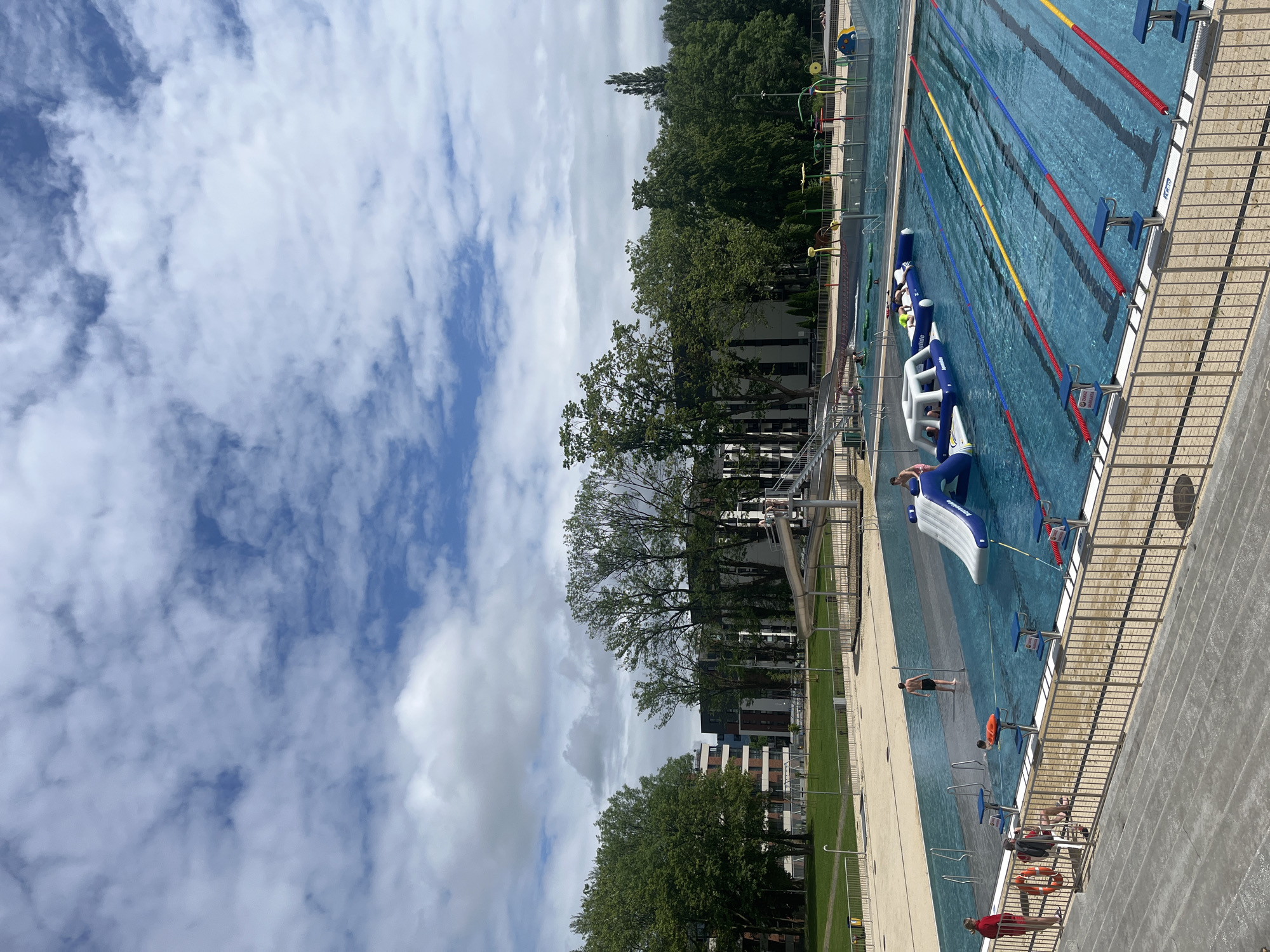 The image shows an outdoor swimming pool complex on a bright, partly cloudy day. The scene is dominated by two distinct pool areas. On the right, there is a large, multi-lane lap pool with blue water, marked by red, blue, and yellow lane lines, and several starting blocks. Text "ARENA" and "MAX LIFE GARANTIE" are partially visible on one of the starting blocks.
Adjacent to the lap pool, on the left, is a leisure pool area featuring large, blue and white inflatable play structures, including a slide and an obstacle course. The brand name "Wibit" is visible on one of these inflatables. Several people, including children and adults, are actively playing on and around these inflatables or swimming in the leisure pool.
Along the concrete deck separating the pools, a few individuals are standing or sitting. At least two people, one wearing a red shirt, appear to be lifeguards, positioned by the pool's edge, with an orange rescue buoy nearby. Another person in dark swimwear stands in the water between the two pool sections.
The pool complex is enclosed by a metal fence and bordered by a strip of green grass. In the background, numerous lush green trees line the horizon, with several modern multi-story buildings visible behind them. The sky is bright blue with scattered white, puffy clouds, indicating good weather for outdoor recreation.