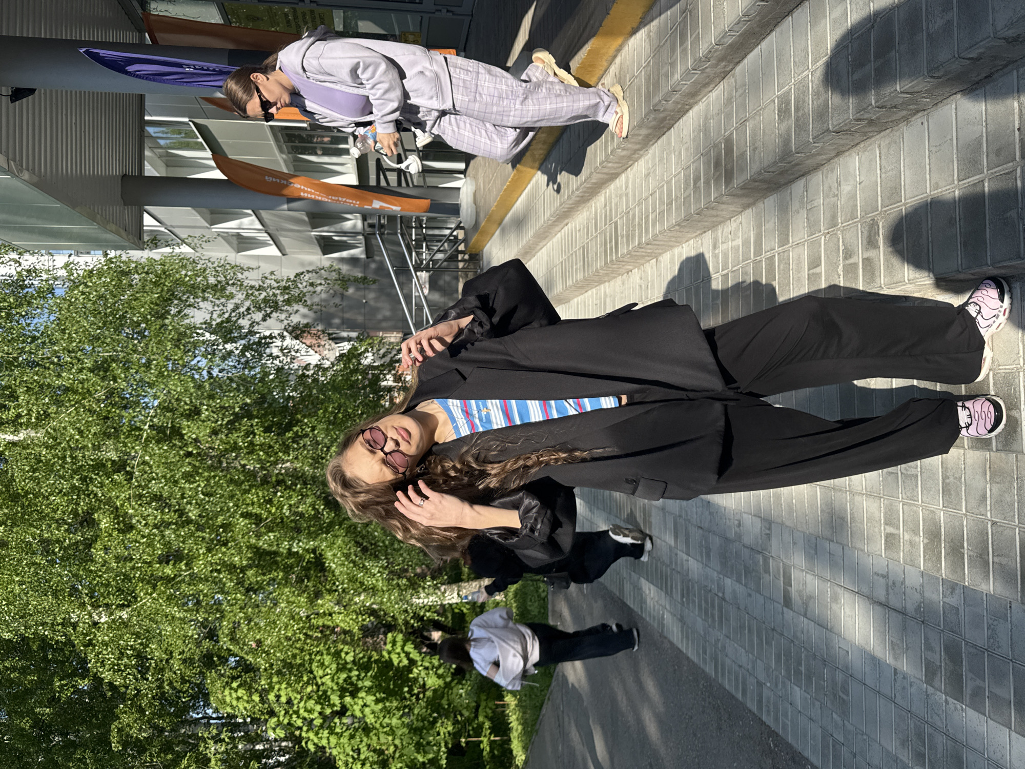 The image shows a daytime scene in Perm, Russia. Two women are present, along with a partial view of a third. The primary woman stands on a sidewalk wearing black pants, a black blazer, a striped shirt, and pink sneakers. She wears round sunglasses and touches her curly hair. A second woman is seen on steps wearing a gray sweatsuit and sandals. A third person is slightly visible in the background. The location seems to be near a building with glass panels and signage. Trees are also visible in the background.