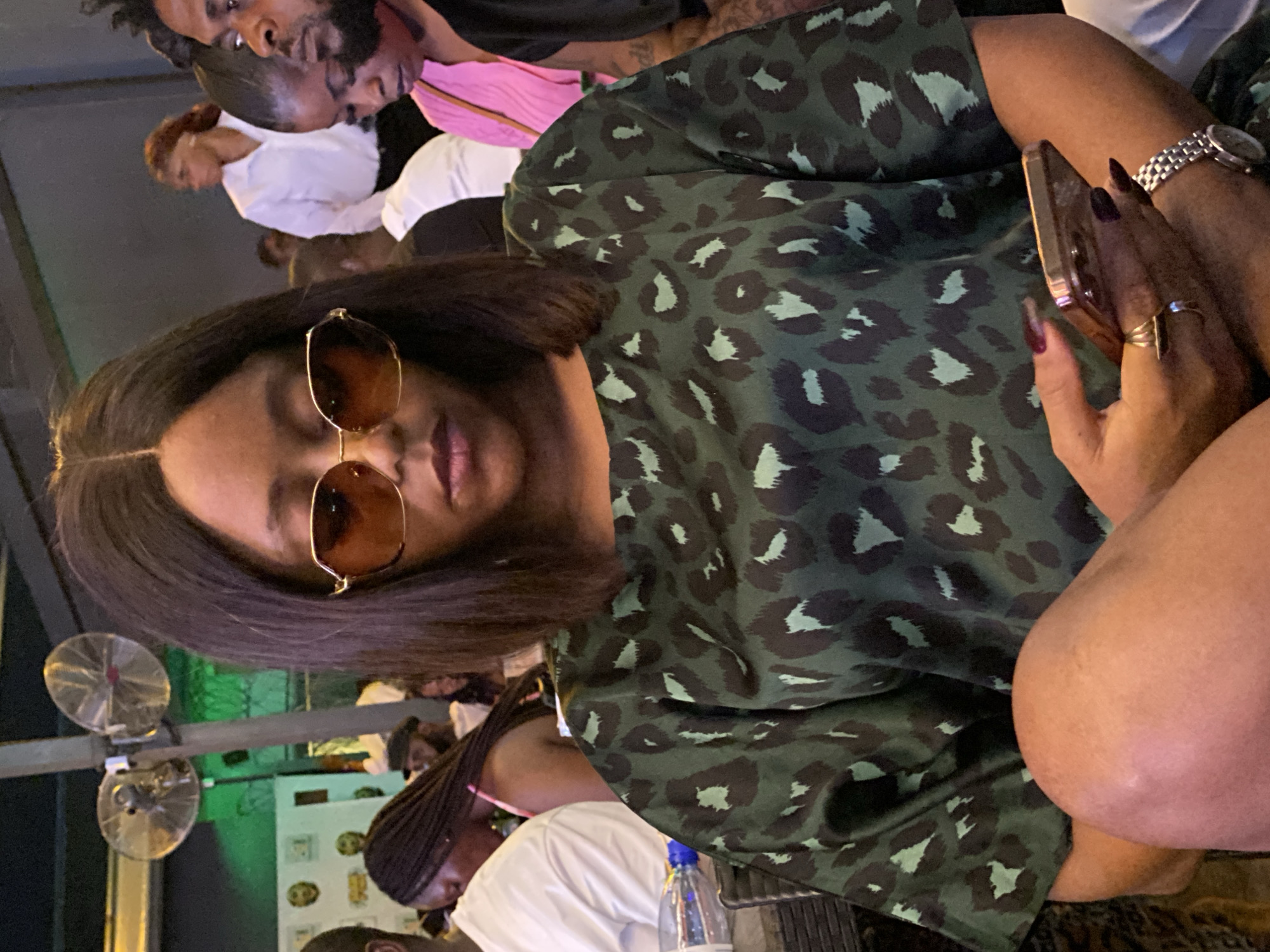 A woman with brown sunglasses and a green and dark leopard print dress is seated indoors. She is holding a cell phone in her right hand. She has long, dark maroon nails and is wearing a silver wristwatch. There are other people blurred in the background. There are electric fans visible in the background. Based on the prompt, the location is Kubwa, Nigeria.