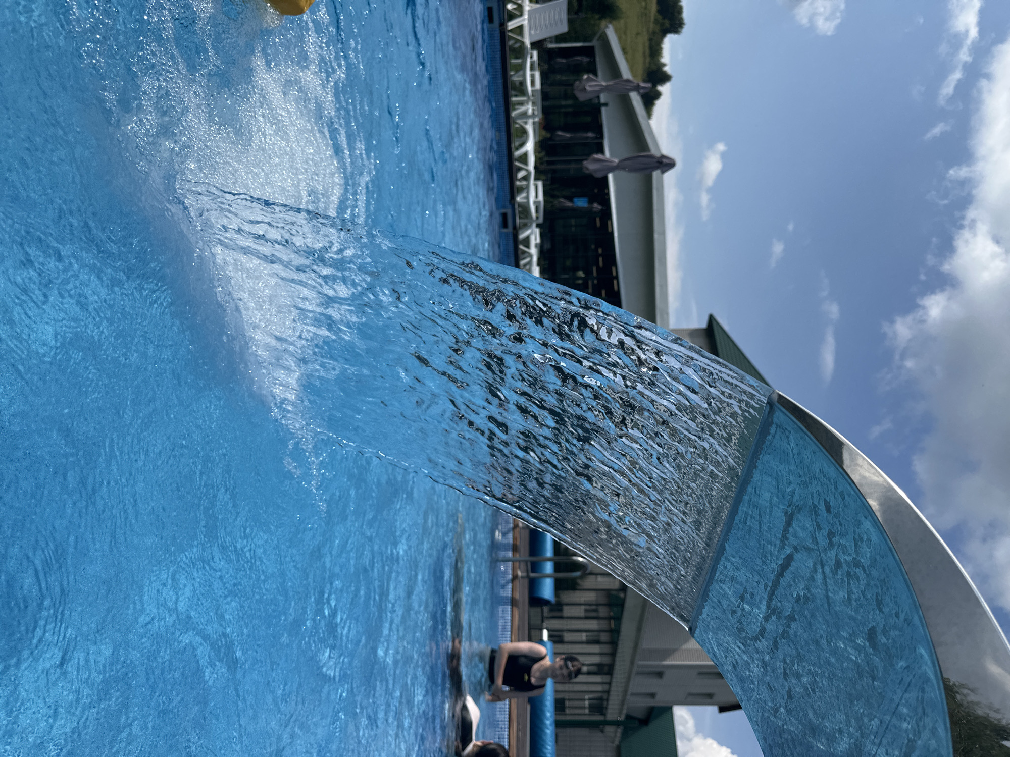 The image captures an outdoor swimming pool on a sunny day. A sheet of water cascades from a curved metal structure, creating a visually dynamic element in the foreground. The pool water is a vibrant blue, reflecting the clear sky dotted with white clouds. In the background, a building with a green roof is visible, alongside lounge chairs and umbrellas, suggesting a resort or recreational facility. Two individuals are partially submerged in the pool, appearing to relax or swim. The overall setting suggests a leisure activity in a warm climate. The location of Даниловка (алтайский), Russia is not ascertainable from the visual information provided in the image.