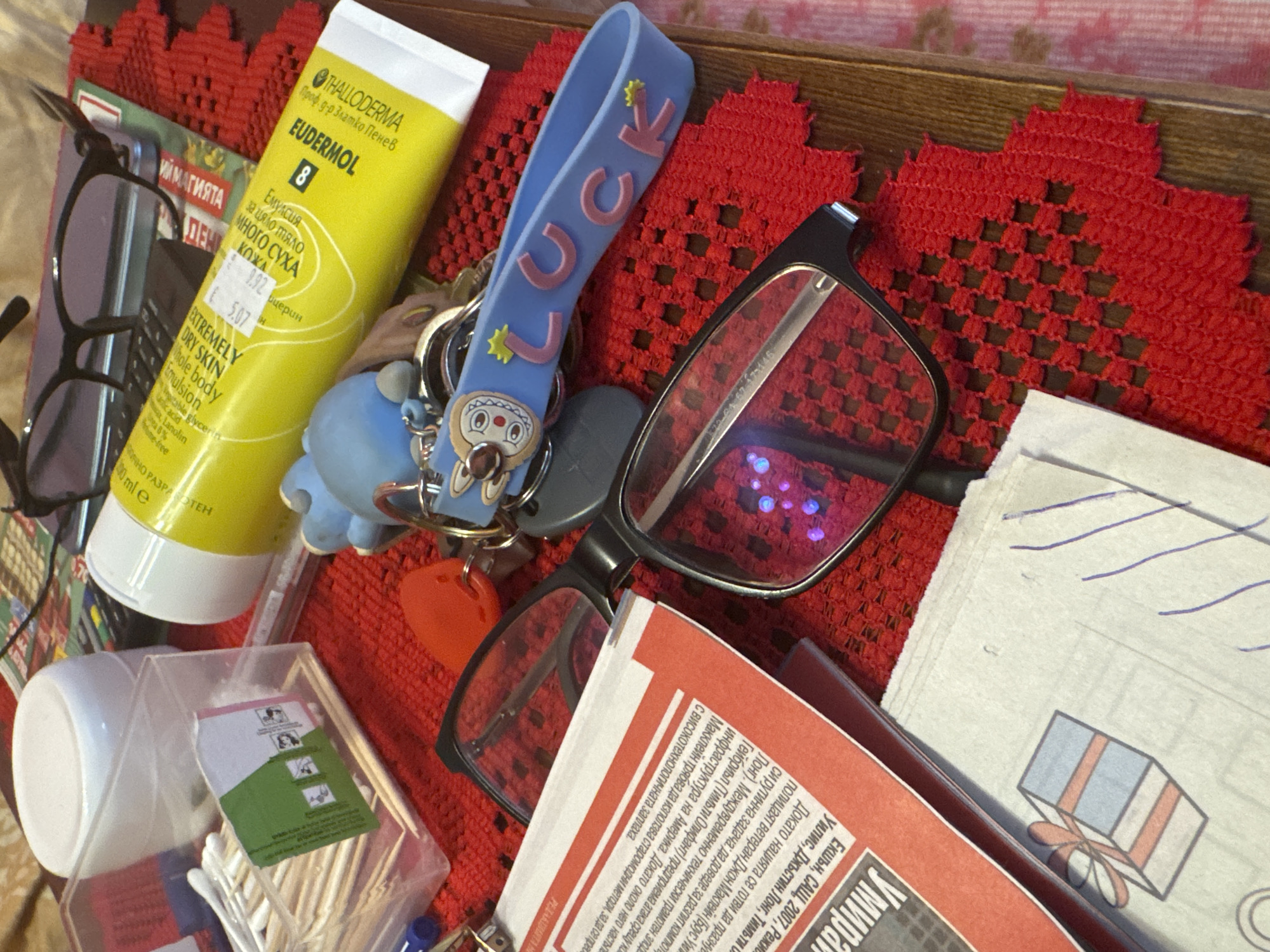 The image presents a cluttered scene, likely an open drawer or box, filled with personal items. Key objects include a tube of "Thalloderma Eudermol 8" lotion with Bulgarian text, glasses, a blue "LUCK" keychain, a box of cotton swabs, a newspaper with Bulgarian text, and a handwritten drawing of a gift. The overall composition suggests a domestic setting. There is no information to suggest the location of Burgas, Bulgaria.