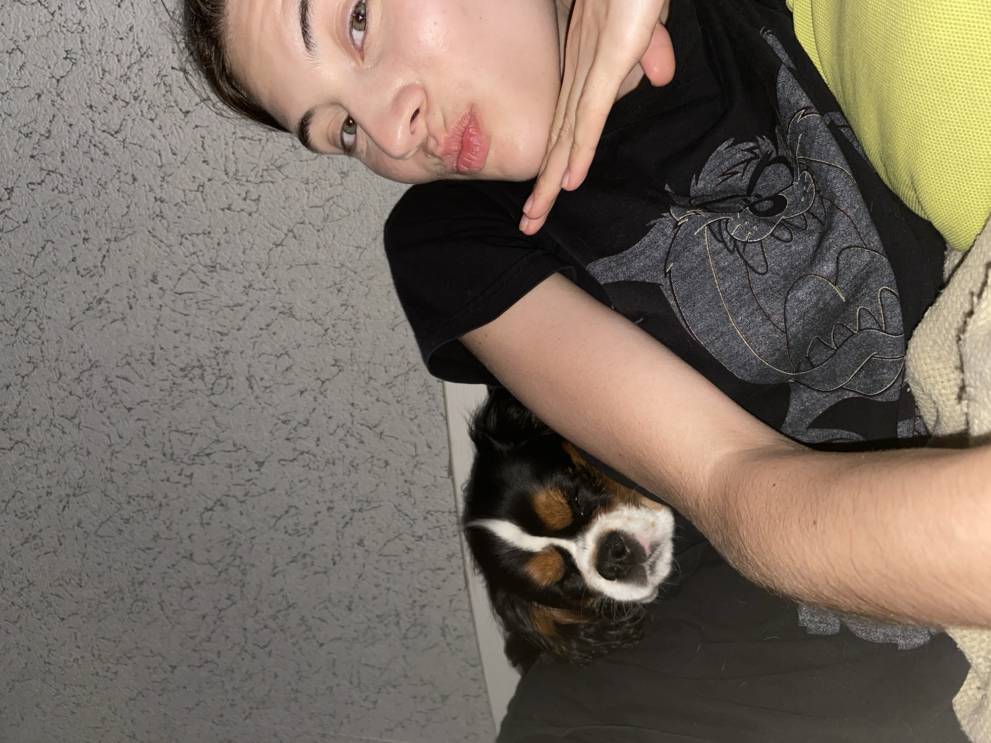 A selfie shows a young person with brown hair and green eyes lying down, with a small black, tan, and white dog nestled beside them. The person is wearing a black t-shirt with a grey cartoon graphic. Their right arm is visible, bent at the elbow and resting on their chest, while their left hand is positioned under their chin. The person is puckering their lips for a kiss, and their eyes are looking towards the camera. The dog appears to be sleeping or resting with its eyes closed. The background consists of a textured grey wall. The scene appears to be indoors, possibly in a bedroom. There is no indication of the location being in Votkinsk, Russia.
