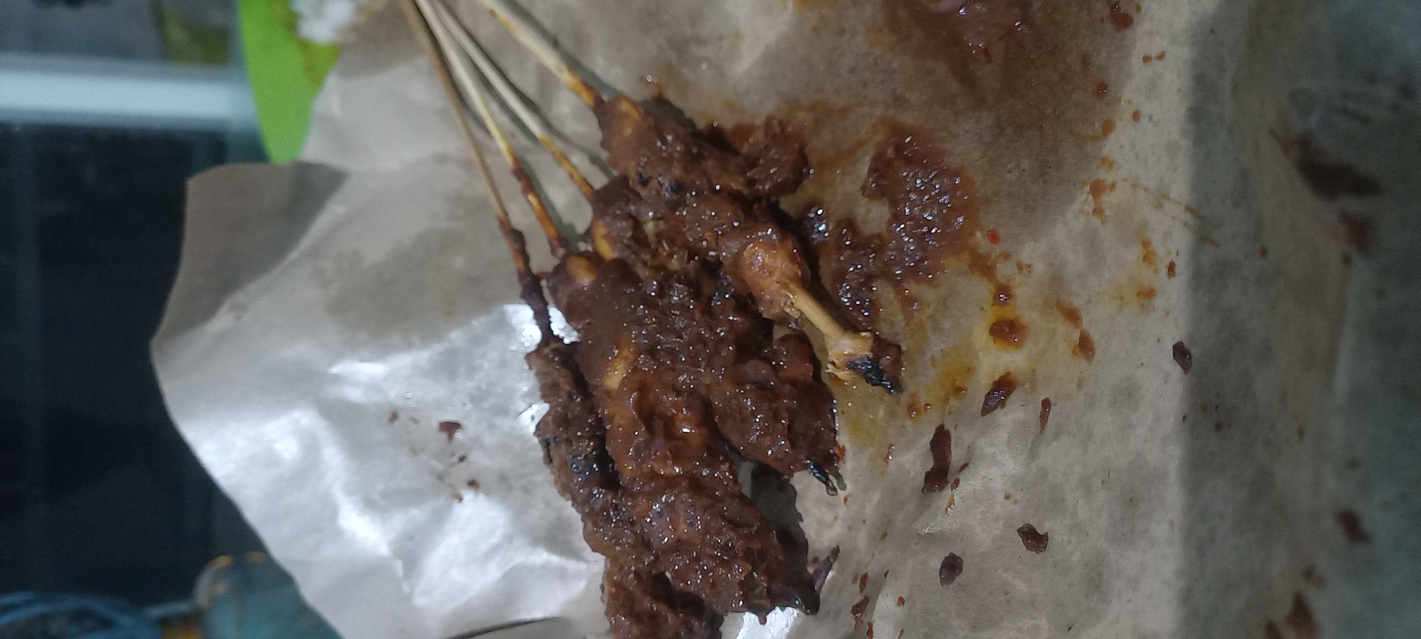 A close-up shot shows several skewers of satay, coated in a dark, glossy sauce, resting on a sheet of brown parchment paper. The satay skewers are arranged diagonally, with the meat pieces appearing to be chicken. Some of the skewers have exposed bone fragments. The parchment paper is creased and stained with the sauce, suggesting it has been used to serve the food. In the background, a blurred, dark area hints at an indoor setting with some faint lighting. The image appears to be taken in Loa Lepu, Indonesia, likely at a food stall or casual dining establishment given the presentation.