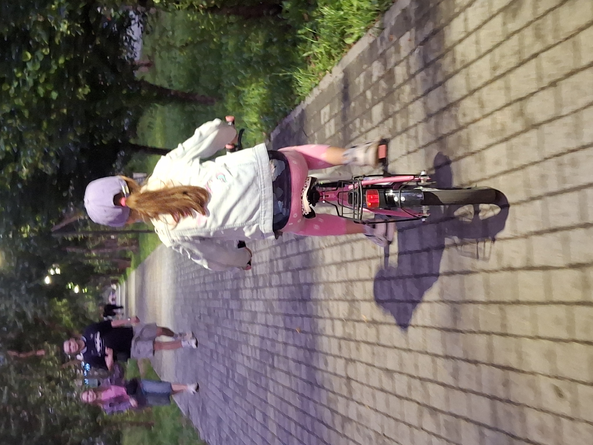 The image shows a child riding a pink bicycle down a paved walkway in a park or recreational area. The child is wearing a white jacket, pink leggings with white polka dots, and a purple cap. Two adults, a man and a woman, are walking on the walkway in the background. The man is wearing a black polo shirt and gray shorts, and the woman is wearing a pink shirt and denim shorts. The walkway is lined with trees and has streetlights visible in the distance. The setting appears to be a city park in Chaikovsky, Russia, likely in the evening or early morning due to the artificial lighting and shadows.