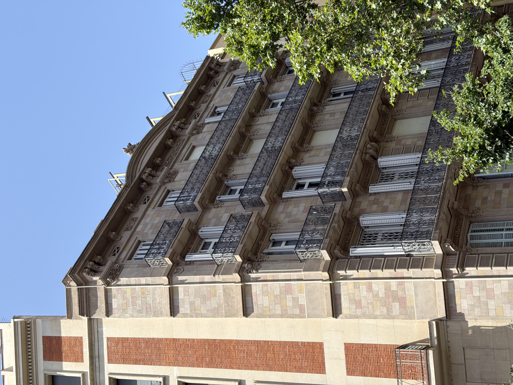 The image shows an exterior view of an ornate, multi-story building in Barcelona, Spain, under a clear blue sky. The building has a facade constructed of light-colored stone with some brick accents. Multiple balconies with intricate wrought-iron railings are visible along the facade, each adorned with decorative elements. The windows have white shutters, some of which are open. A large, leafy green tree partially obscures the right side of the building. The architecture suggests an older, possibly historic building common in European cities. There are no people or vehicles visible in the image.