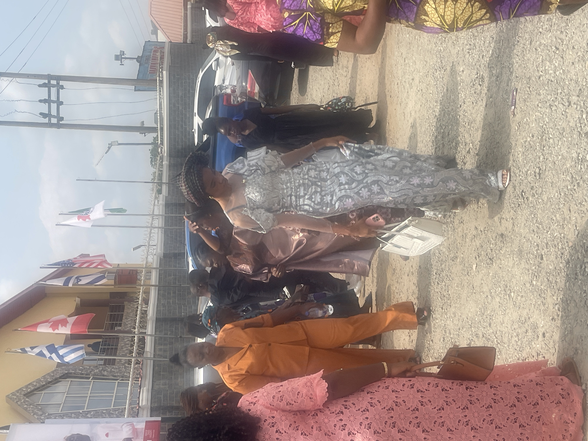 A group of people, mostly women, stand in front of a building with several flags of different countries. The women are wearing a variety of colorful dresses and suits. Some of them are holding purses or bags. The flags include those of Nigeria, the USA, Israel, Canada, and Greece. The location is likely Abuja, Nigeria, based on the flags and the clothing of the people.