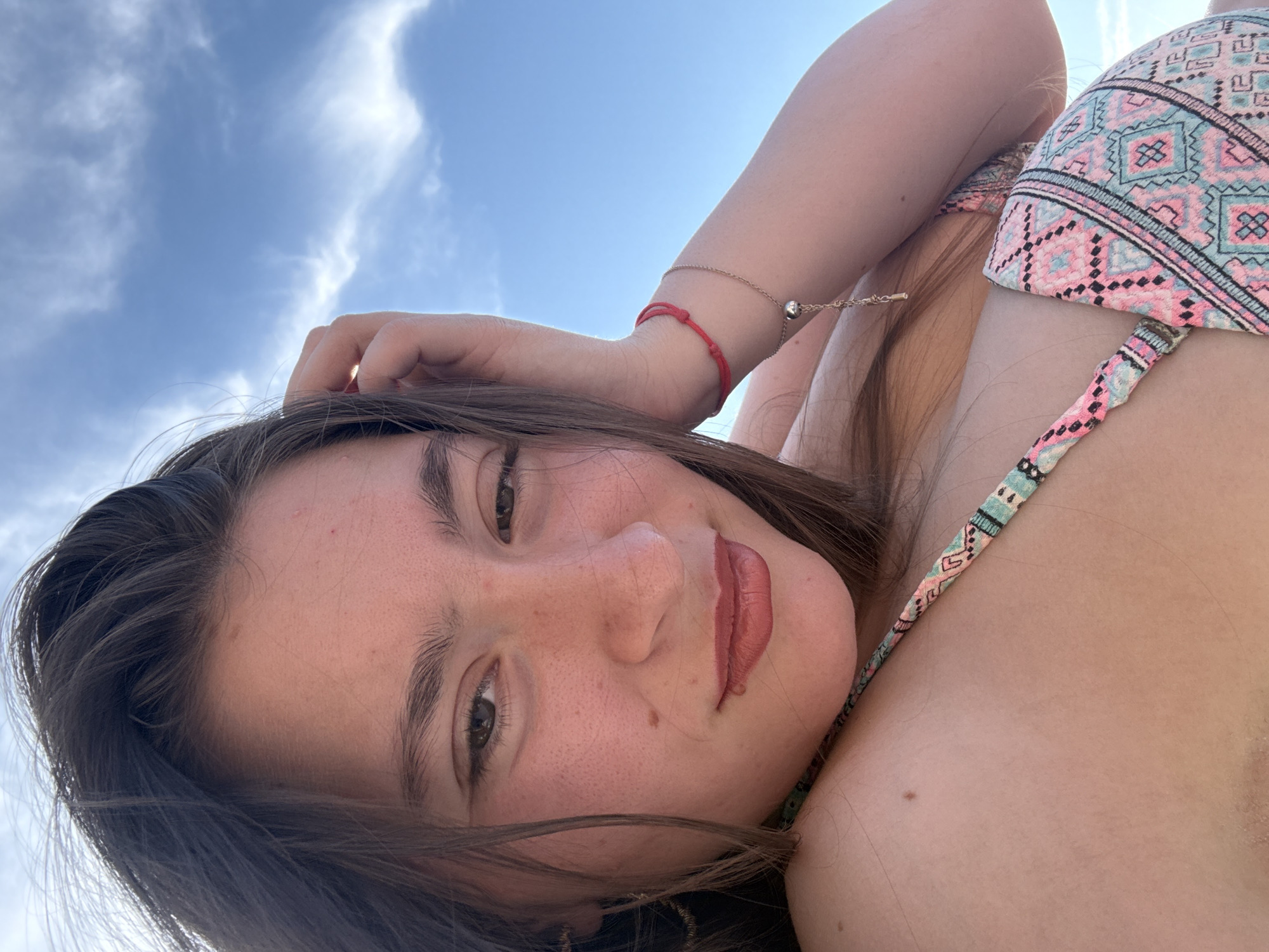 The image shows a young woman lying down outdoors, with a blue sky and clouds visible behind her. She is wearing a colorful bikini top with a geometric pattern in shades of pink, blue, and white. A thin gold bracelet with a small silver charm adorns her wrist, as well as a red string bracelet. Her hair is dark brown and falls around her face, which is tilted upwards. She appears to be relaxed and possibly enjoying a sunny day. The overall lighting suggests it is daytime, and the presence of clouds indicates the weather is partly cloudy. The location context suggests this photo was taken in Sozopol, Bulgaria.
