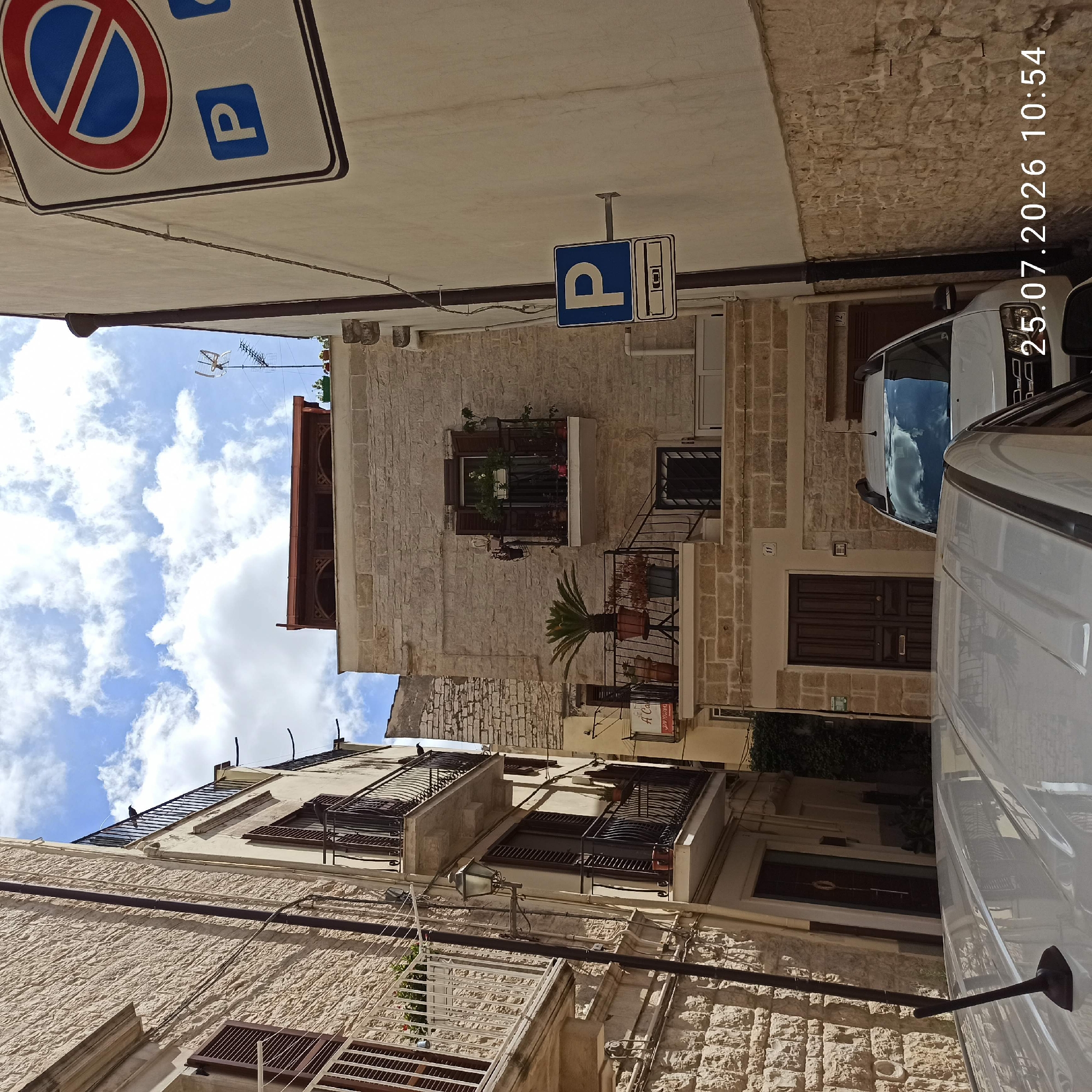 This is a street-level view of a narrow alleyway in Ruvo di Puglia, Italy, with buildings constructed of light-colored stone on both sides. The sky above is partly cloudy.
On the left, a multi-story building features balconies with ornate black metal railings and brown shutters on the windows. One balcony has a white railing and a planter with greenery. A lamppost is attached to the building's facade.
The building on the right is also made of stone and has a balcony with black railings. A potted palm tree and other plants are visible on a lower balcony. Above this, a second-floor balcony has shuttered windows and more plants. A small sign that reads "A CA" is visible below this balcony.
In the background, a white SUV is parked in the alleyway. The image is overlaid with a timestamp: "25.07.2026 10:54".
Traffic signs are visible on the right wall, including a "no parking" sign with a blue circle and red cross over a blue 'P', and a blue 'P' sign indicating parking, with a secondary sign beneath showing a silhouette of a car and a horizontal line.
The overall impression is of an older, well-maintained urban environment.