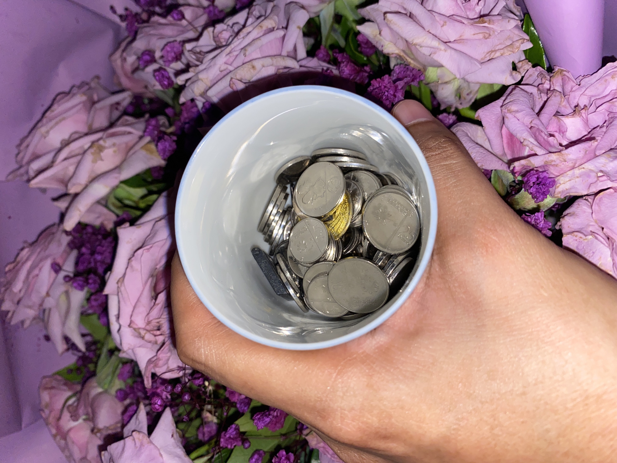 A person's hand holds a small, white ceramic cup filled with Philippine coins against a backdrop of pink and purple roses and small purple flowers. The coins are primarily silver in color, with one noticeable gold coin. The image was taken indoors. The location is Davao City, Philippines (based on the coins).