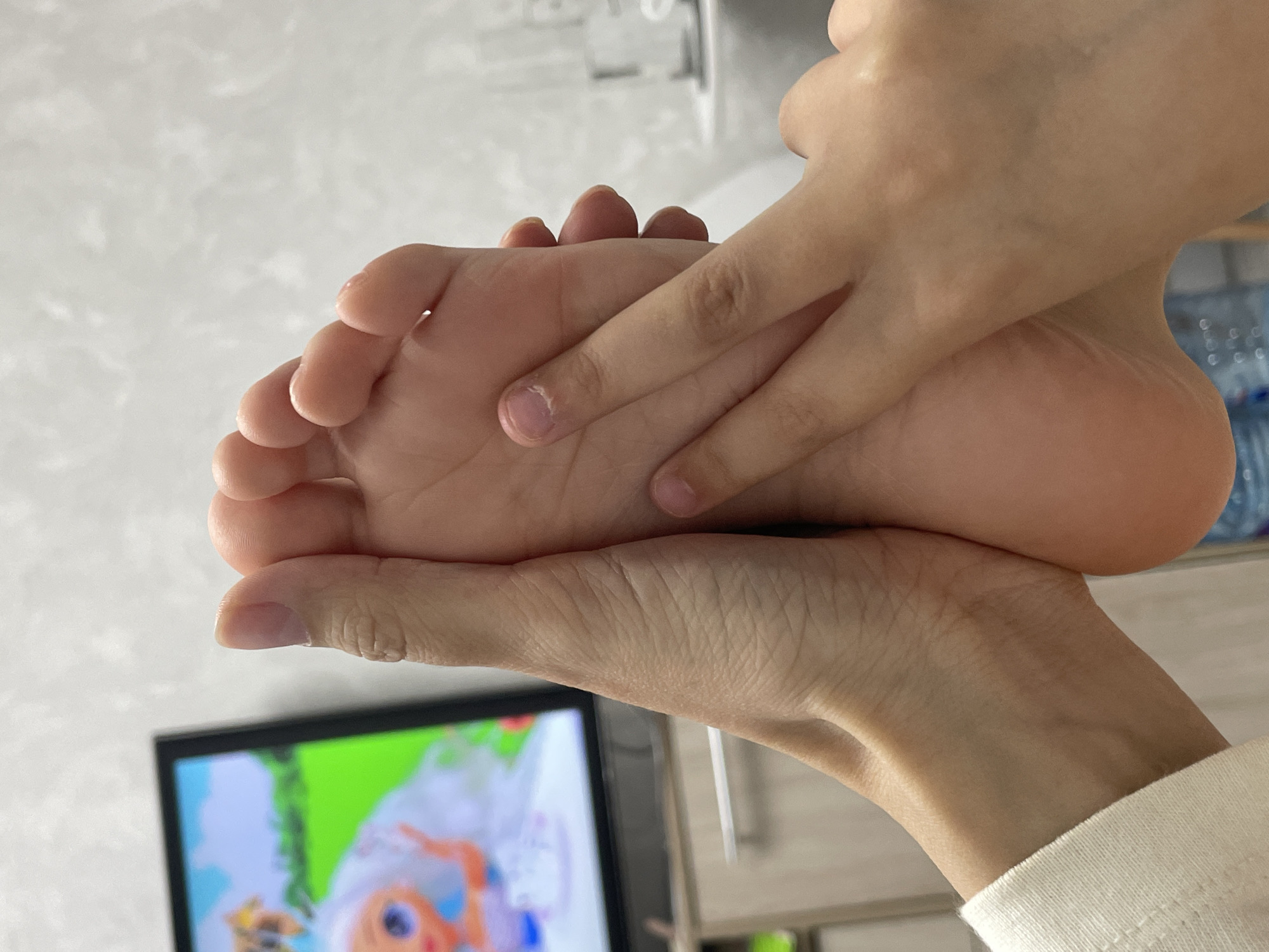 A close-up, indoor shot captures a person's hands gently holding their own bare right foot. The hands, appearing to belong to a single individual, are positioned to cup and potentially massage the sole and toes. The skin tone is light, and the fingernails are natural and neatly trimmed.
In the blurred background, a television screen is visible in the lower left, displaying a brightly colored cartoon or animated program. The screen shows a character, possibly a doll, with white hair or a wig against a prominent green and blue background. In the upper right background, out-of-focus clear plastic bottles or containers are stacked on a shelf or counter. A light-colored, possibly white or cream, fabric sleeve is visible on the right edge of the frame.
The setting is an informal indoor space, consistent with a domestic environment, located in Izhevsk, Russia. The specific time of day or weather conditions are not discernible from the image. The activity suggests a moment of relaxation, self-care, or examination. There is no visible text present in the media file.