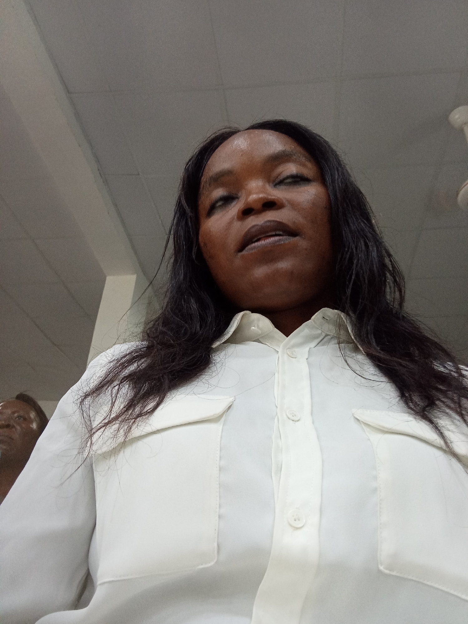 A low-angle, medium shot captures a Black woman from the chest up, looking upwards and to her right. She has long, dark, wavy hair that falls around her shoulders and chest. She is wearing a loose-fitting, white collared shirt with two chest pockets and white buttons down the front. Her facial features are partially obscured by the angle, but she has dark eyes and her mouth is slightly open. In the background, on the left side of the frame, a person's head and shoulders are visible from a distance, appearing to be looking at the camera. The background consists of a textured ceiling with visible light fixtures and what appears to be a fan. The overall lighting is bright, suggesting an indoor environment. The scene is likely a public space or a residential interior in Kason, Nigeria.