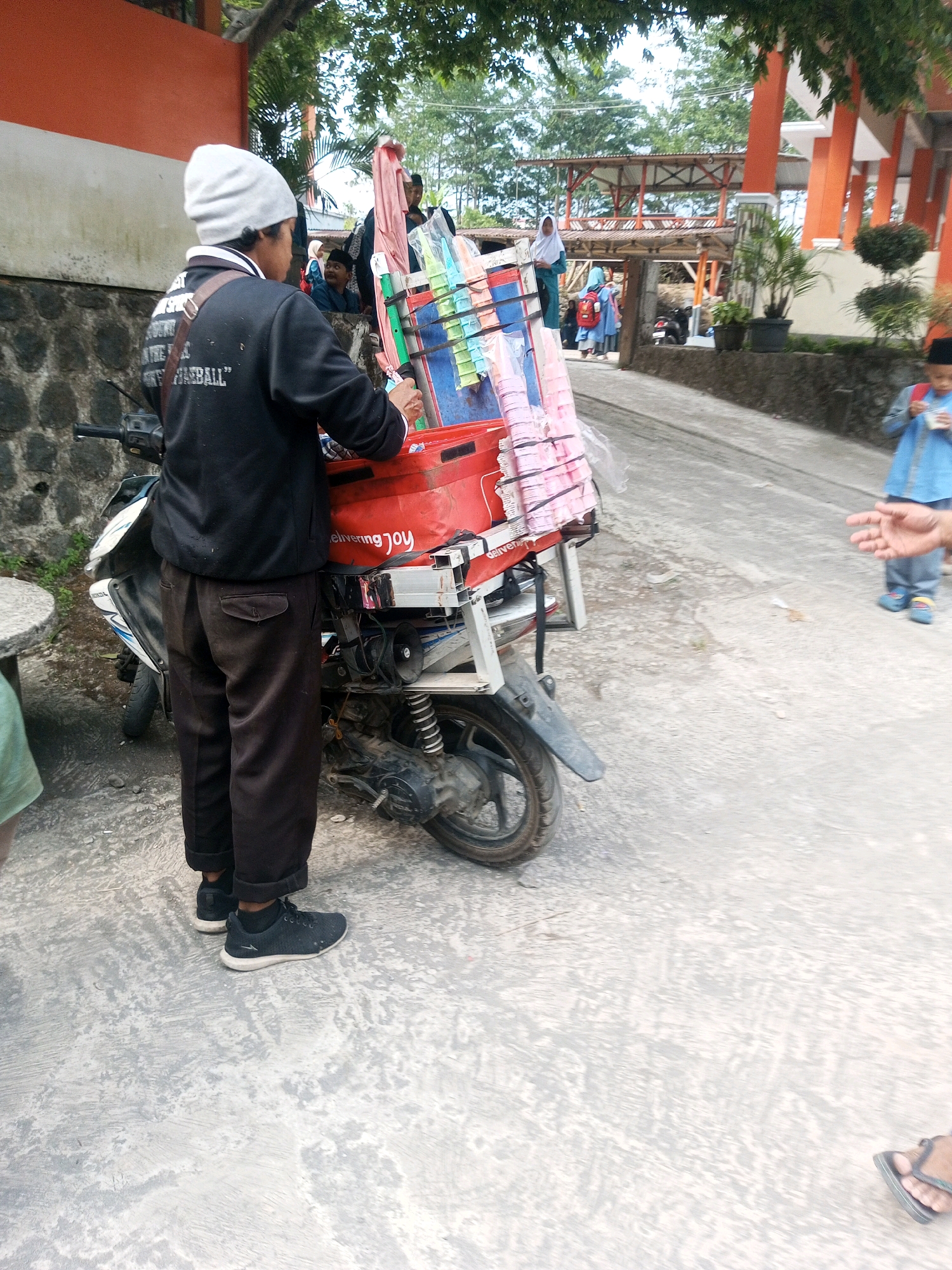 In Kalibeber, Indonesia, a person wearing a gray beanie and a black jacket stands next to a motorcycle with a red box containing colorful items on the back. The person is wearing dark pants and black athletic shoes. The motorcycle is parked on a paved surface, possibly a street or walkway, with a wall and vegetation in the background. A young boy in a blue shirt and pants is visible in the distance, with his hand extended. The scene appears to be outdoors, with trees and buildings suggesting an urban or semi-urban environment.