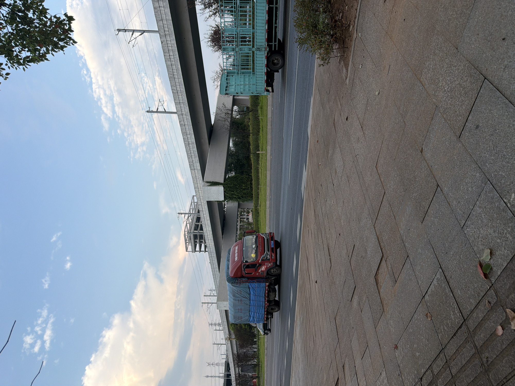 A red truck with a blue tarp is traveling on a multi-lane road in Jiangjin, China. The truck is in the right lane of two visible lanes of traffic. To the right of the red truck, a light blue truck with a metal cage bed is also traveling in the right lane. The road is bordered by a sidewalk made of grey paving stones. In the background, an elevated concrete structure, possibly a bridge or overpass, crosses the frame. Overhead power lines and poles are visible above the elevated structure. The sky is blue with scattered white clouds, and a tree with green and red leaves is visible in the upper right corner. The overall scene depicts urban transportation infrastructure under a partly cloudy sky.