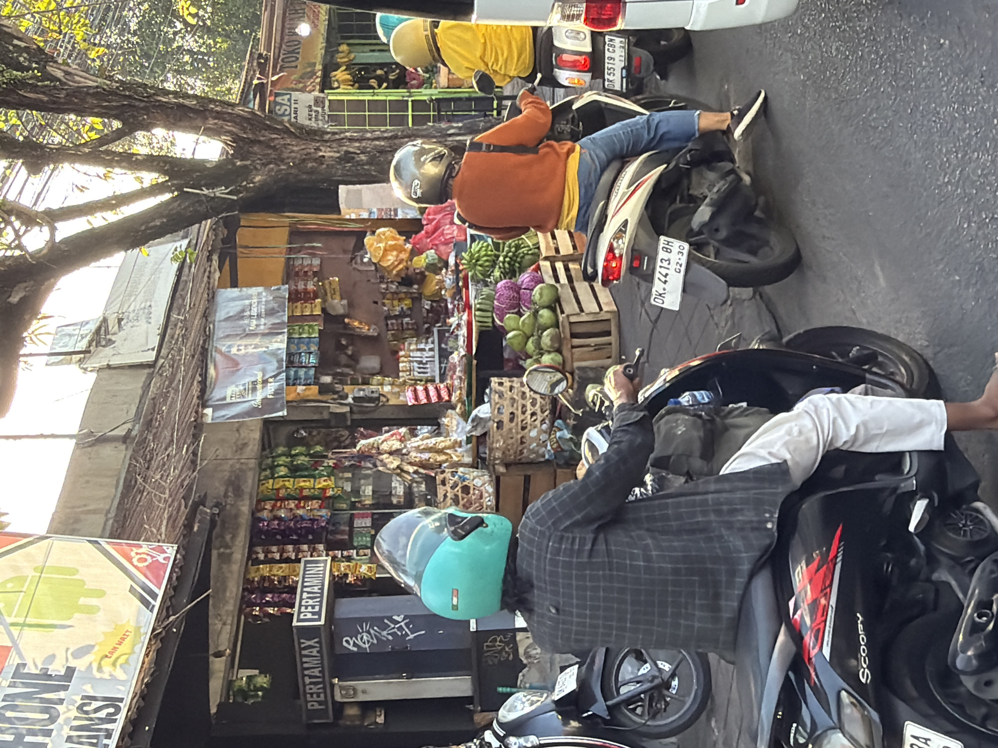 The image depicts a street scene in North Kuta, Indonesia, likely during the day, with sunlight filtering through trees. In the foreground, two individuals are riding motorcycles. The person on the left is wearing a checkered shirt and a turquoise helmet, riding a black scooter with "Scoopy" branding. The person on the right is wearing an orange sweater over a yellow shirt and blue jeans, with a silver helmet, riding a white scooter. Behind the motorcycles, there are several small storefronts or stalls selling various goods, including packaged snacks and produce like coconuts and bananas. Signs in Indonesian are visible, with "PERTAMAX PERTAMINI" prominently displayed on one stall, indicating a fuel station or convenience store. The overall environment suggests a bustling commercial area with traffic and pedestrian activity.