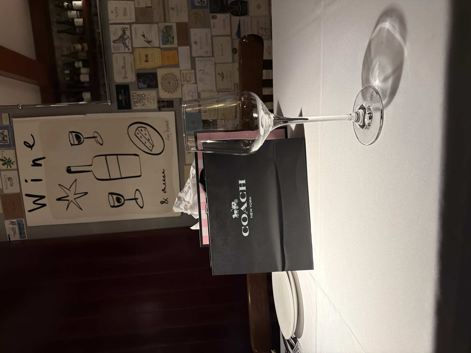 The image depicts a close-up of a table setting with a wine glass and a Coach shopping bag. The wine glass is clear and empty, with a long stem and a wide bowl, casting a shadow on the white tablecloth. Beside the wine glass is a black Coach shopping bag with the brand's logo and "NEW YORK" printed in white. The bag appears to be partially open, revealing some pink tissue paper inside.
To the left of the wine glass, a portion of a framed poster or artwork is visible. It features a minimalist illustration of a wine bottle, a wine glass, a starburst, and a wedge of cheese. The word "Wine" is prominently displayed in large, bold lettering.
The background shows shelves filled with wine bottles and various labels, suggesting a wine cellar or a restaurant setting. There are also scattered papers and small framed items on the wall behind the wine bottles, possibly decorative elements or wine labels. The overall scene is indoors, likely in a dimly lit environment, with the primary light source creating reflections on the wine glass and the white tablecloth. The presence of the Coach bag and the wine-themed artwork suggests a celebratory or gifting occasion. The location context is Qinhuai District, China. No people are visible in the image.